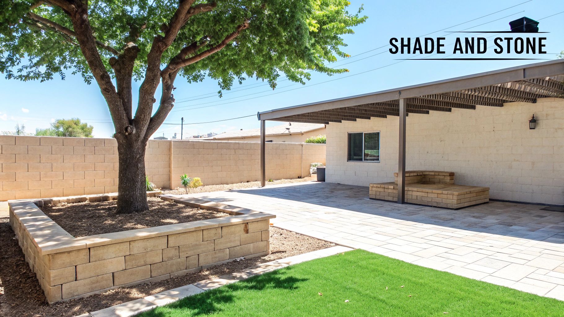 A well-maintained backyard featuring a large shade tree in a stone planter, a paved patio, a built-in stone bench, and a small patch of green grass.
