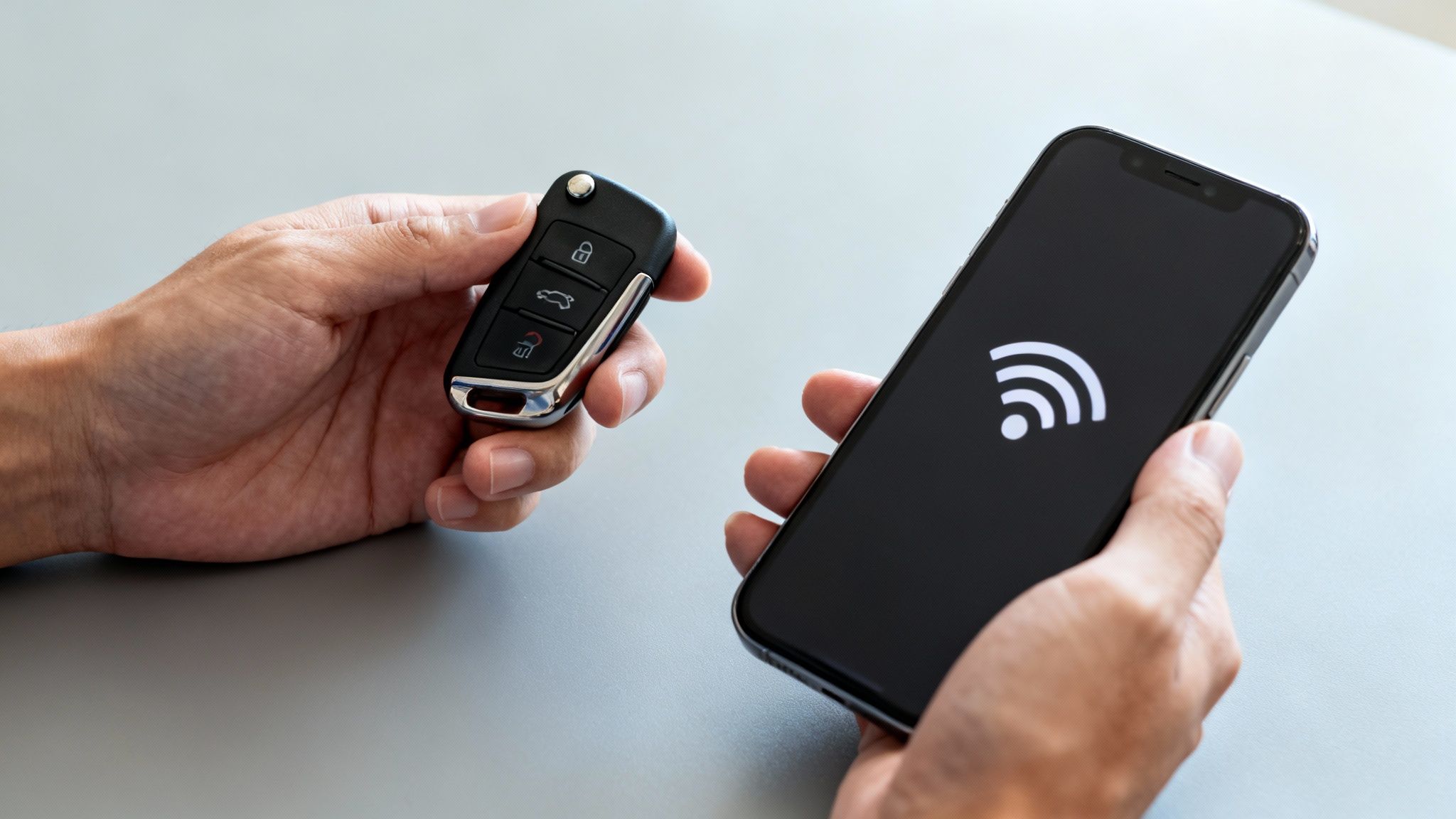 Hands holding a car key fob and a smartphone displaying a wireless signal symbol, suggesting connectivity.
