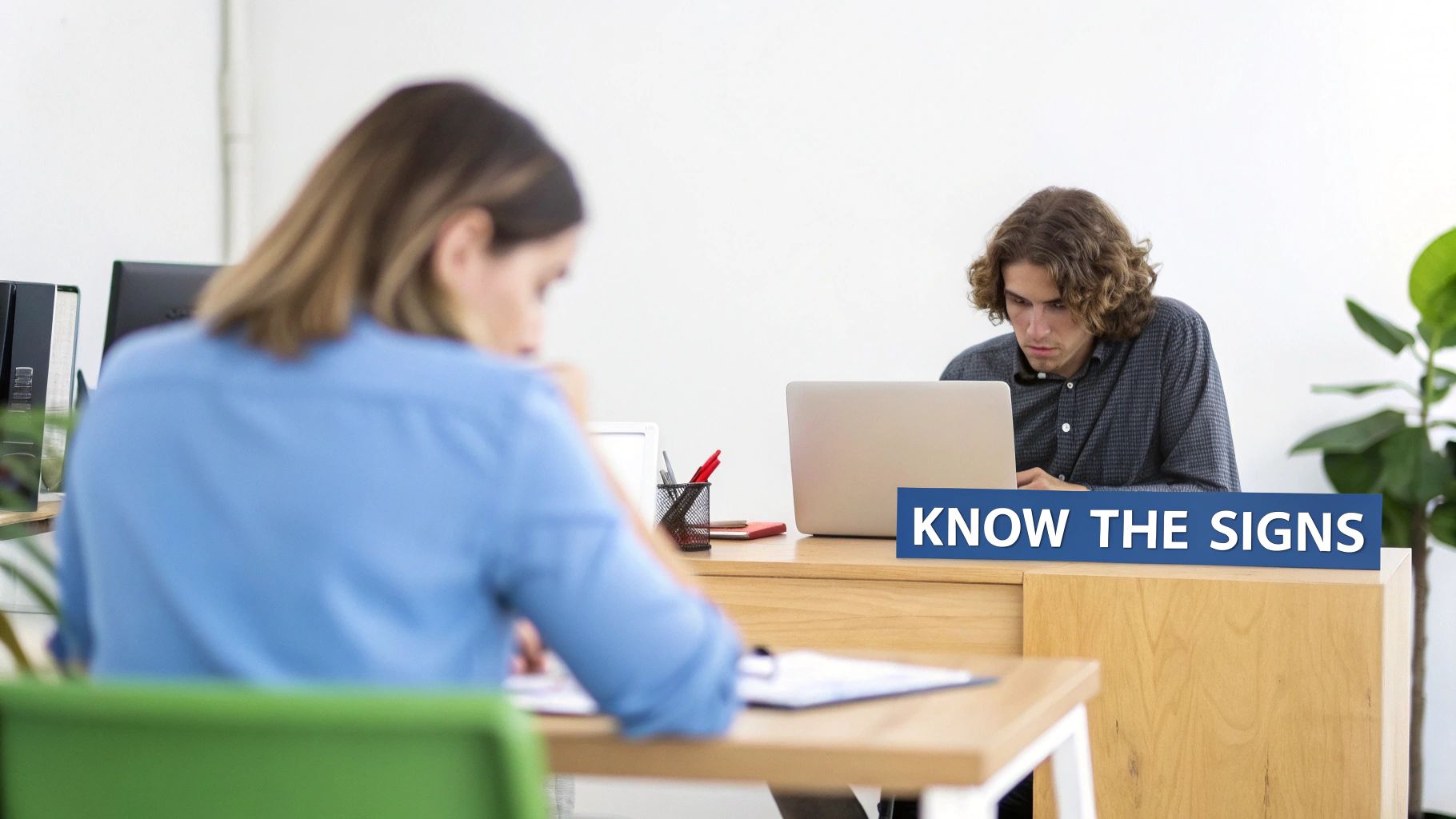 Two people working in a bright office, one focused on a laptop, with a "KNOW THE SIGNS" banner.