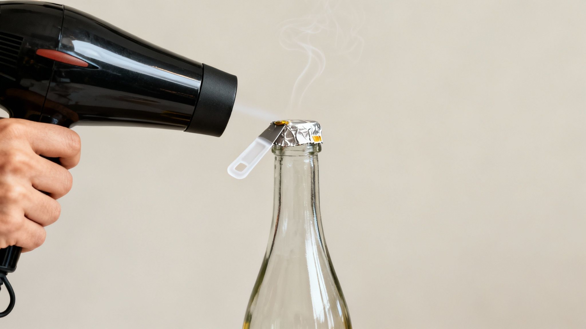 Close-up of a hand holding a hairdryer heating a foil-wrapped bottle top, with smoke rising.