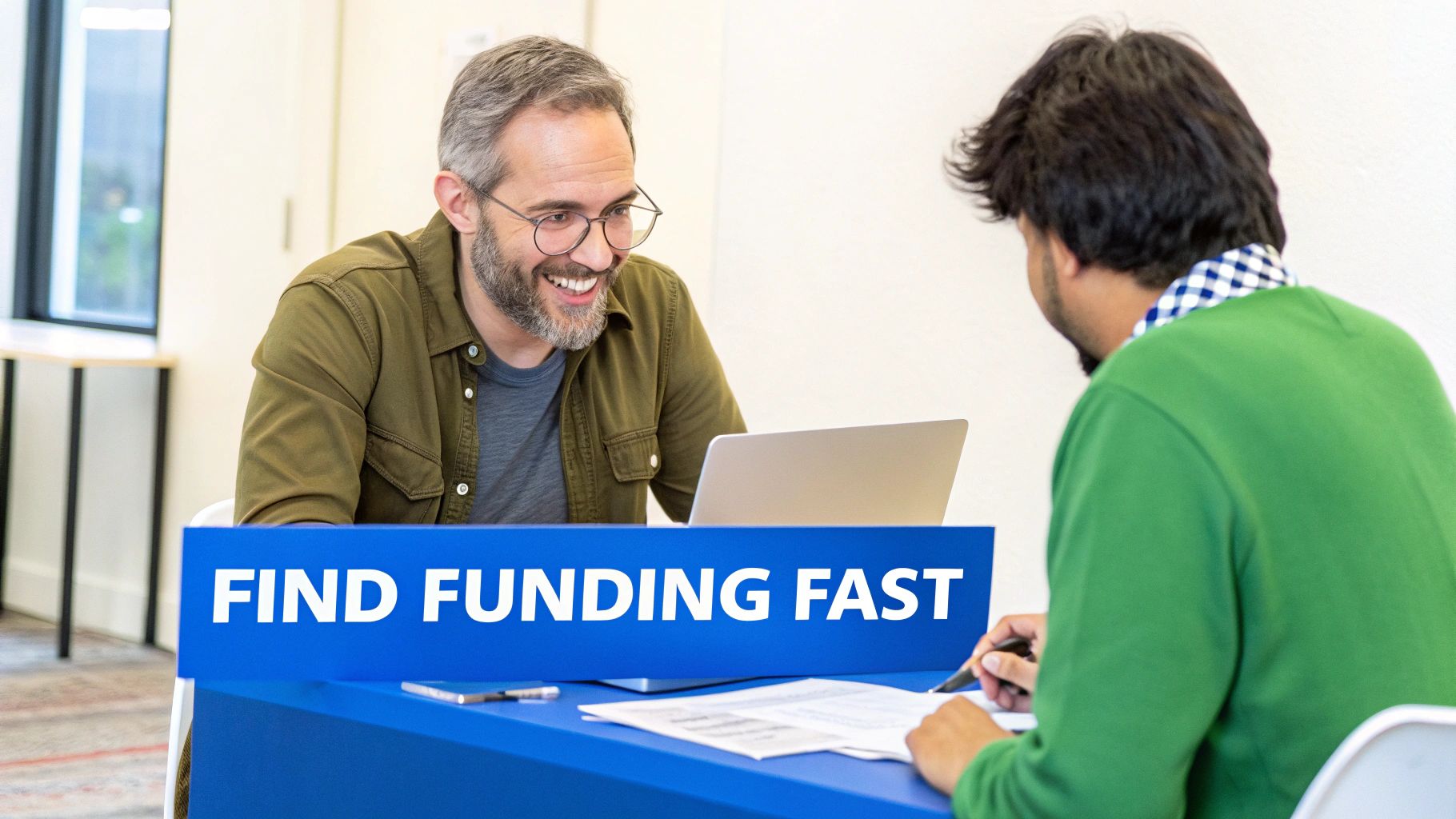 Two smiling men, one with glasses, discussing documents at a blue table with a laptop and a 'FIND FUNDING FAST' sign.