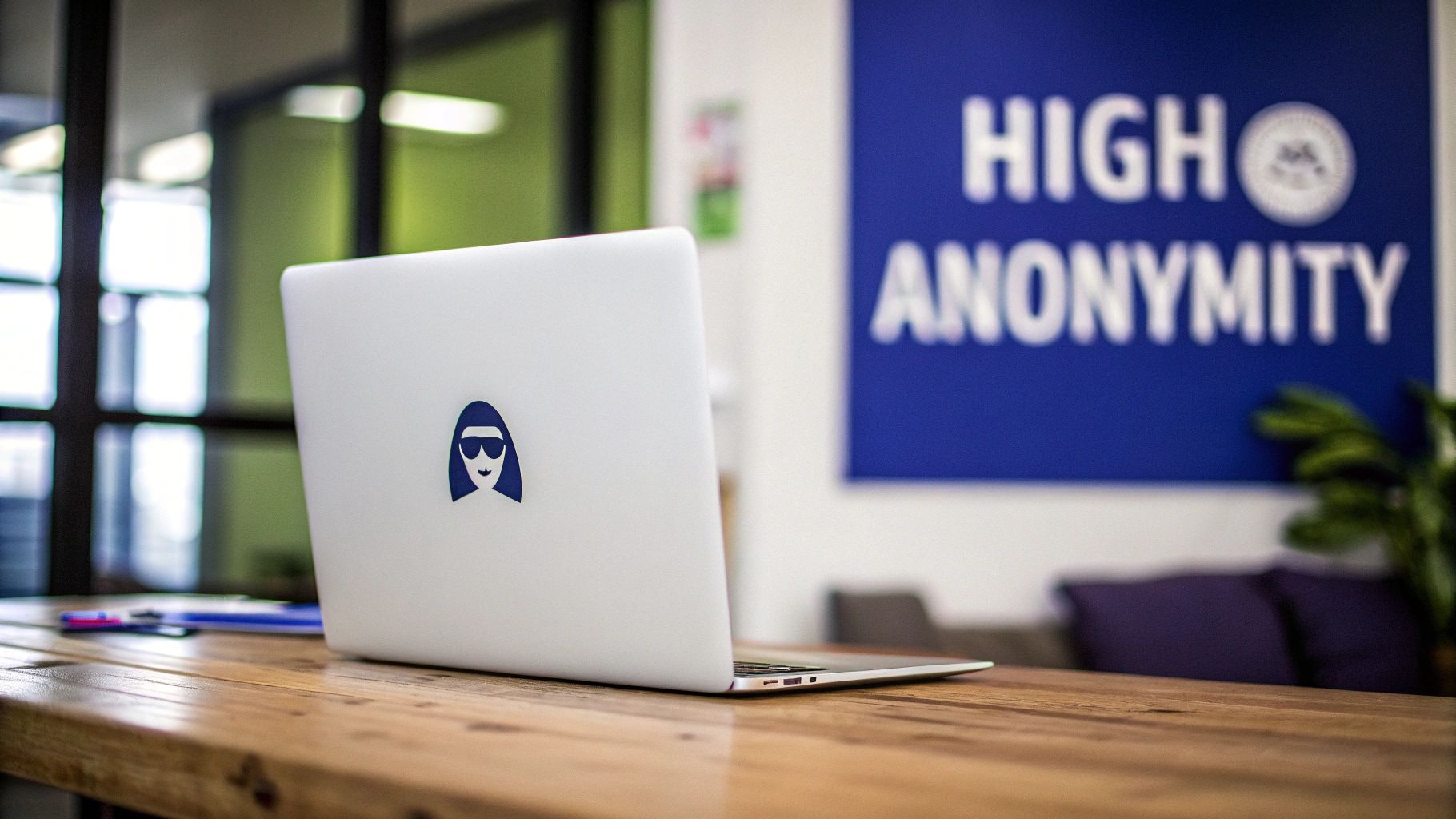 A white laptop with a blue person-in-sunglasses logo on a wooden desk, in front of a blurry 'HIGH ANONYMITY' sign.