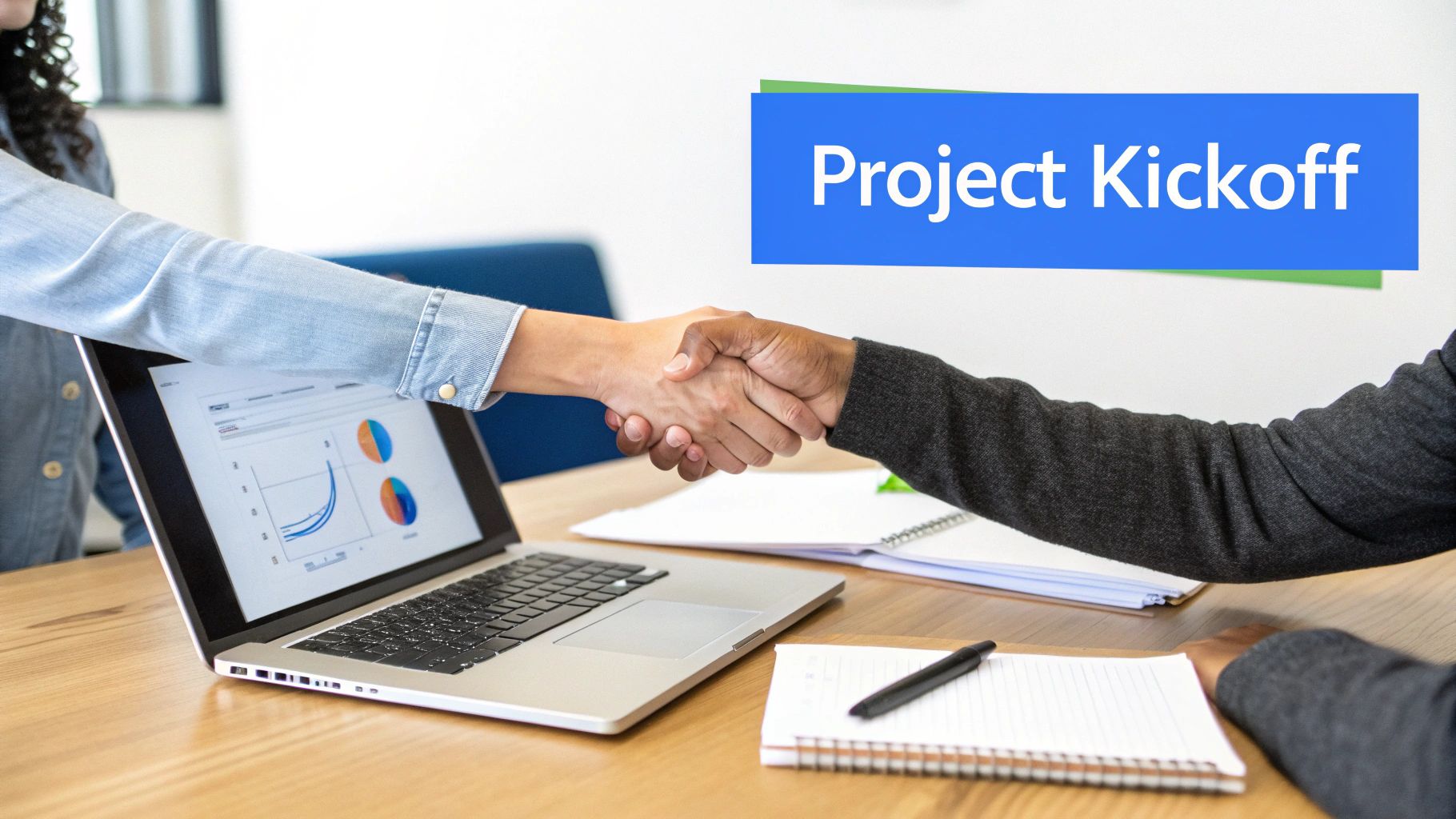 Two diverse individuals shake hands over a laptop displaying project charts during a kickoff meeting.