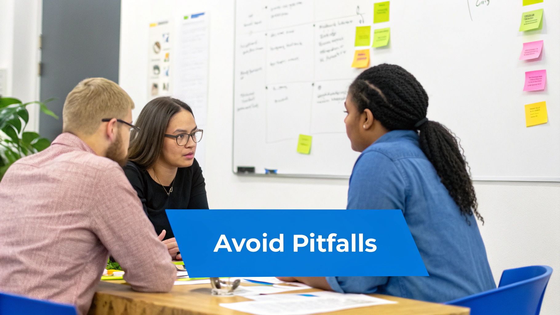 Three diverse colleagues engaging in a discussion around a table with a whiteboard full of sticky notes in the background.