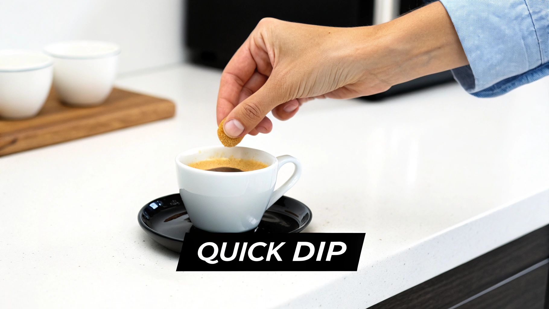 A hand dips a small, round biscuit into a fresh cup of espresso on a white counter.