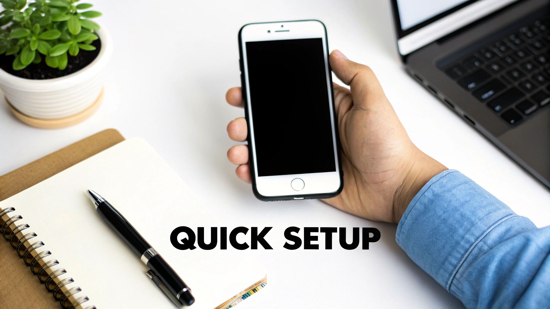 A hand holding a smartphone with 'QUICK SETUP' text, alongside a laptop, notebook, and plant on a desk.
