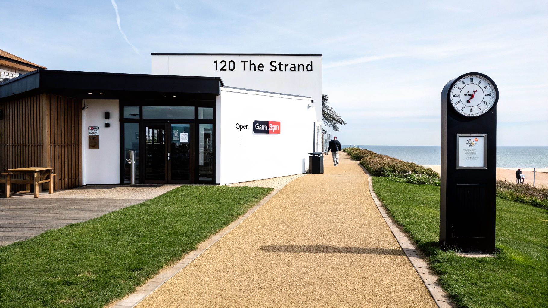 Modern cafe building '120 The Strand' by the beach, with a clock tower and walking path.