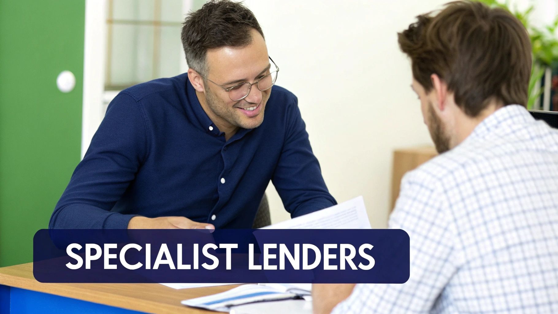 A smiling man in glasses discusses documents with a client at a desk, a specialist lenders meeting.