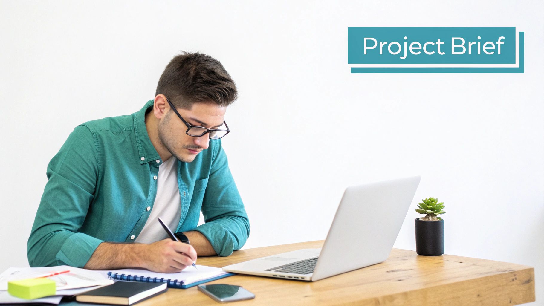 A man in glasses, wearing a green shirt, writing in a notebook at a desk with a laptop, working on a project brief.
