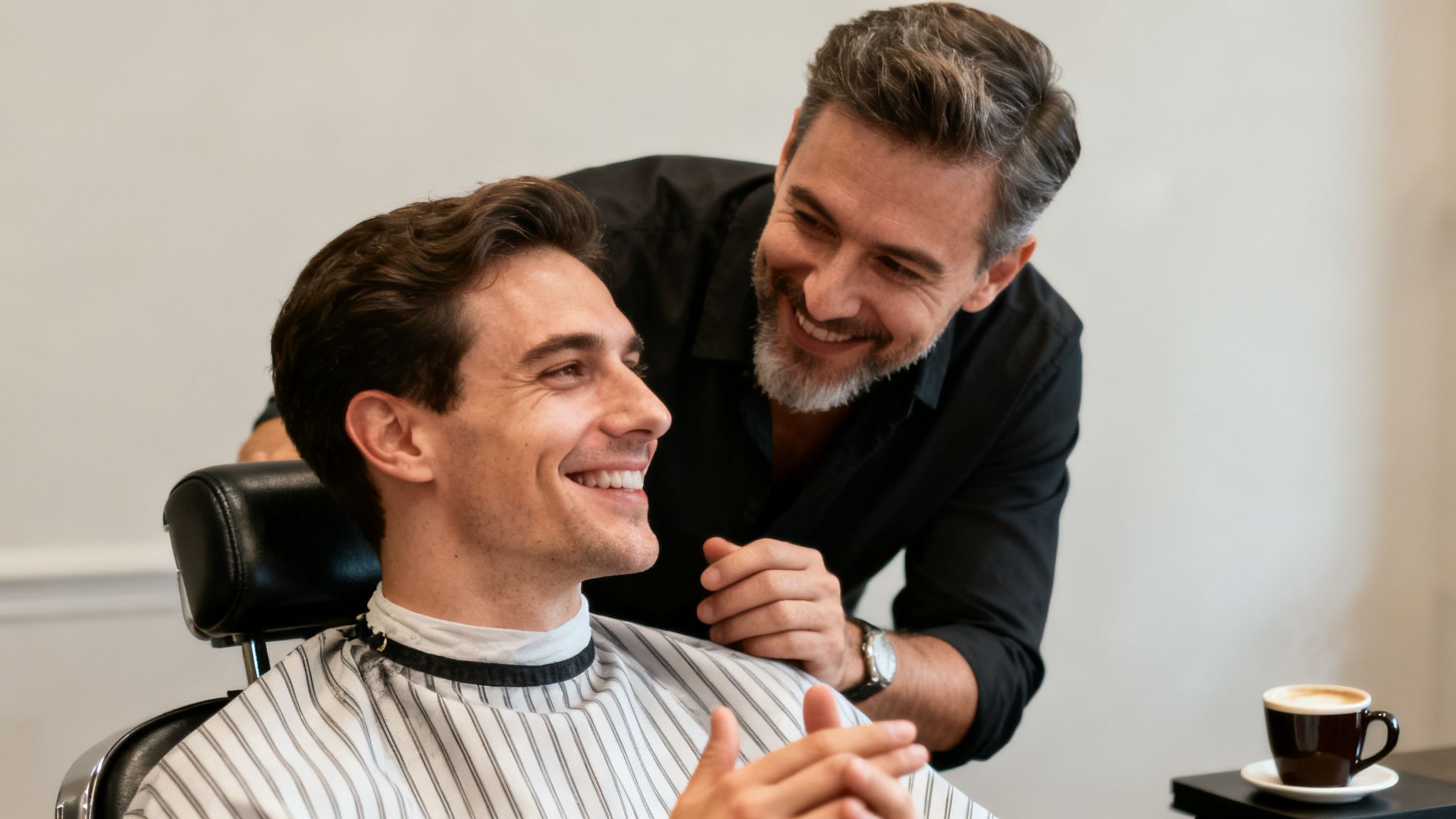 A smiling barber with a grey beard looks at his happy client in a barber chair.