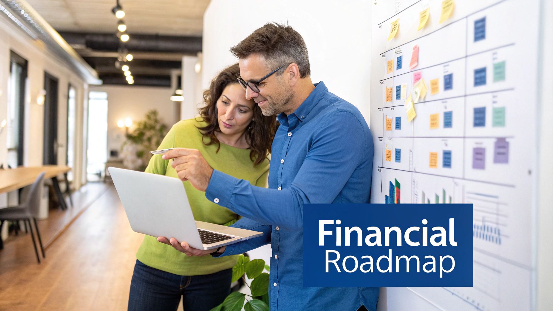 Two colleagues review financial data on a laptop, discussing a financial roadmap with a planning board in the background.