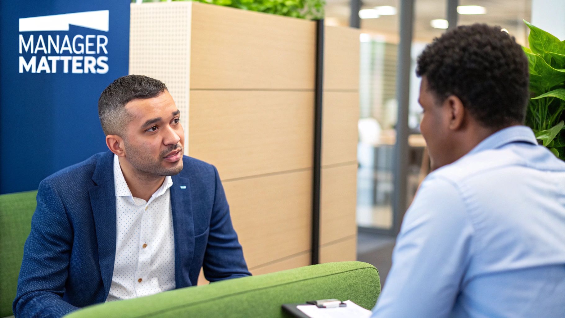 Two professionals discussing in an office with a 'Manager Matters' sign, focusing on the speaker.