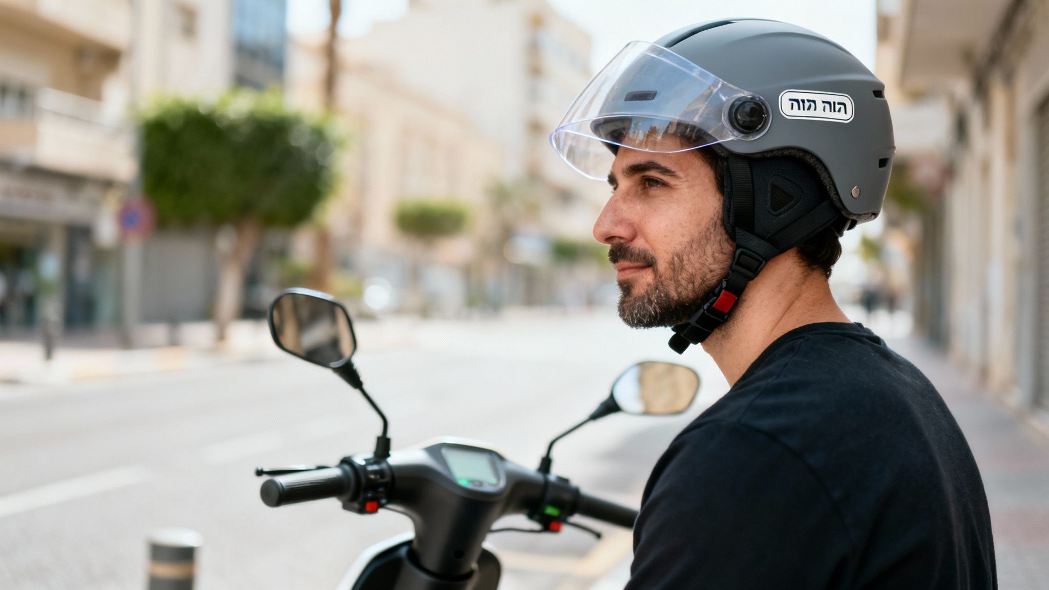 גבר עם זקן קצר חובש קסדה אפורה עם משקף, יושב על קטנוע חשמלי בעיר.