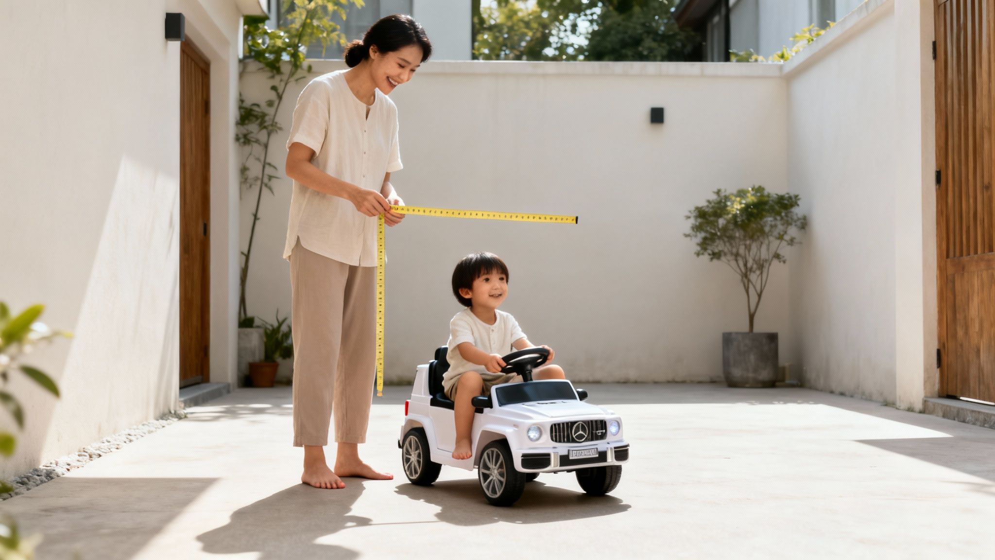 אישה מחייכת מודדת מכונית צעצוע לבנה עם סרט מדידה צהוב, בעוד ילד קטן יושב בה בחצר שטופת שמש.