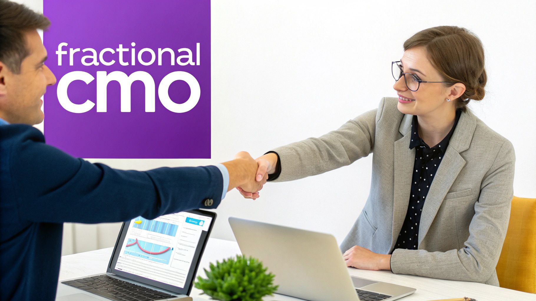 Two business professionals shake hands in an office with laptops and a 'fractional CMO' sign.