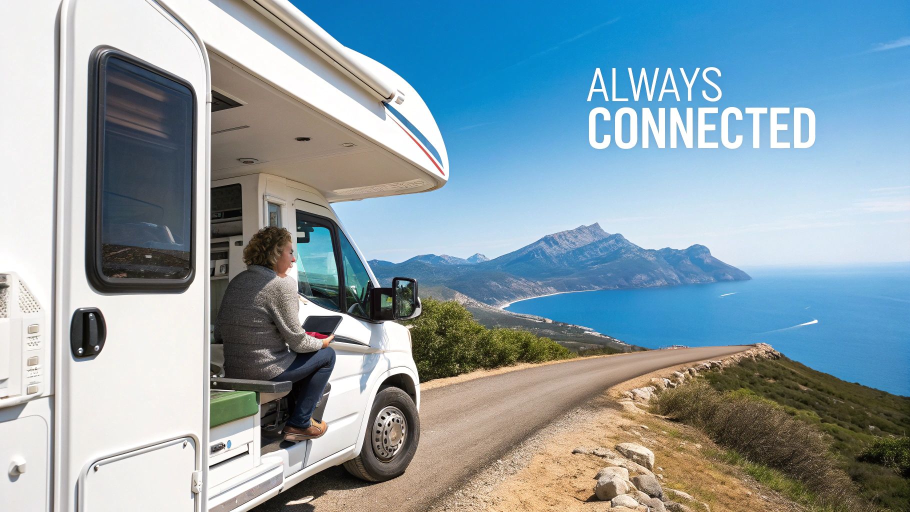 A person uses a tablet in a white RV overlooking a scenic coastal view with mountains.