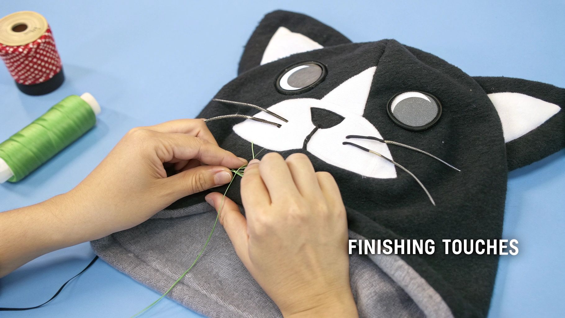 Hands sewing green thread onto a black and white cat costume, adding finishing touches.