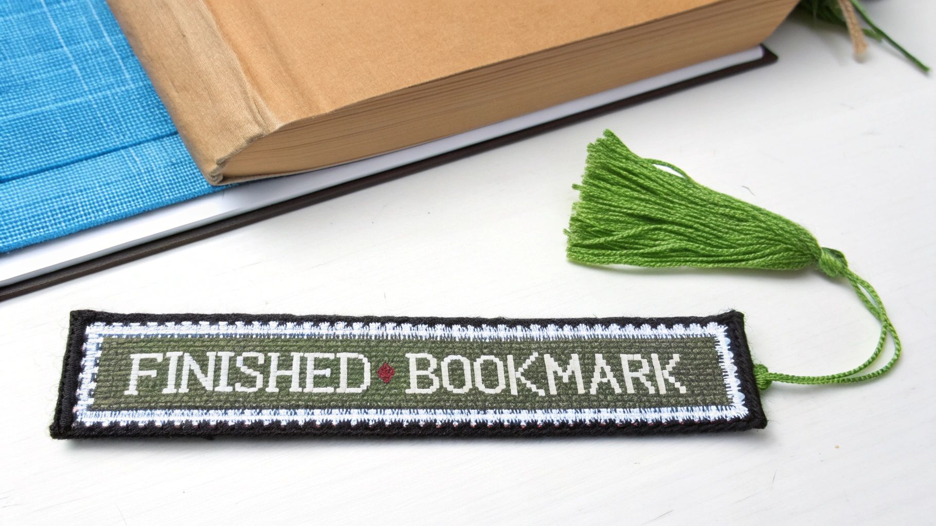 A handmade cross-stitch bookmark with 'FINISHED BOOKMARK' text and a green tassel next to books.