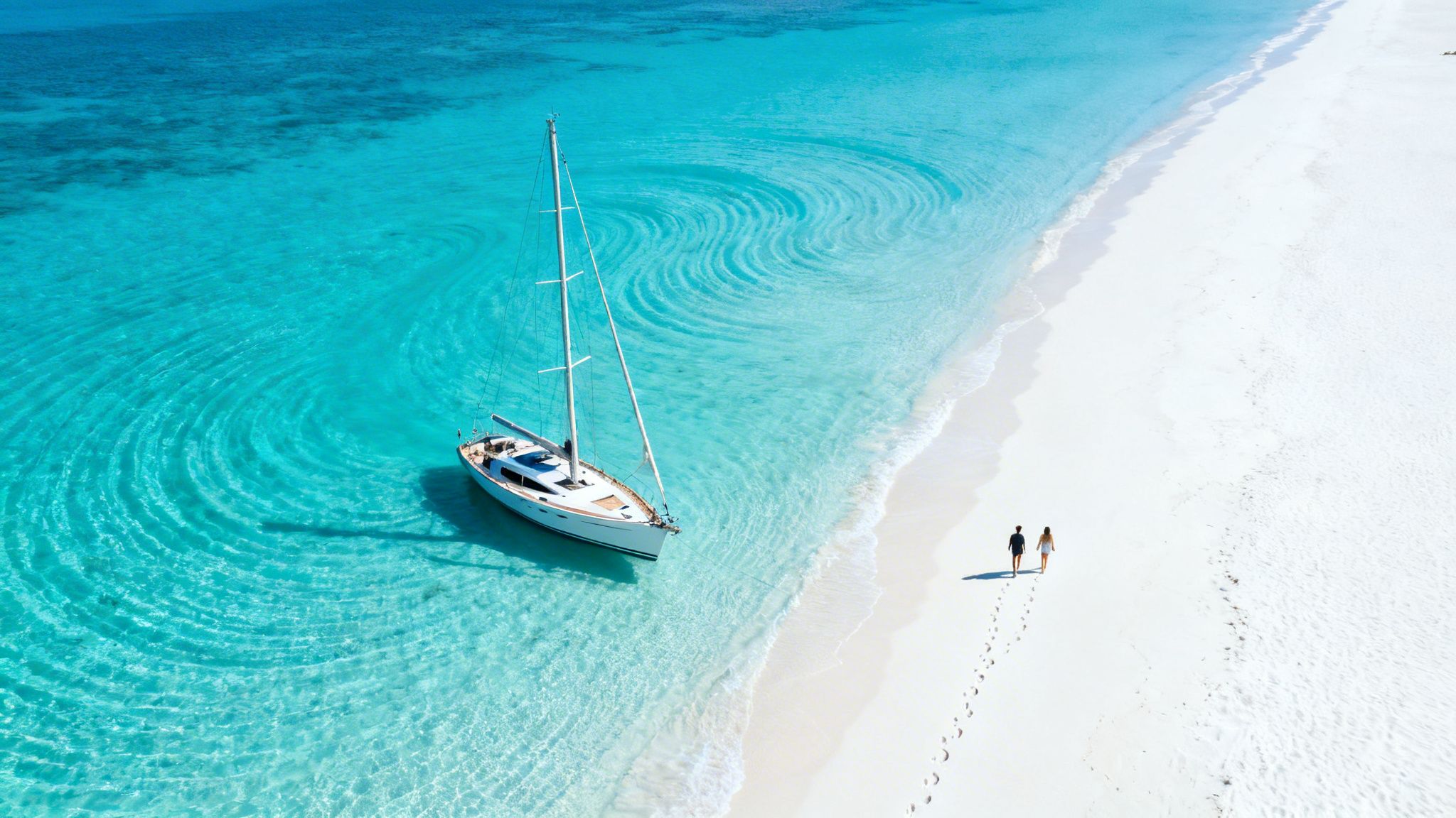 Aerial view of a white sailboat in turquoise water next to a pristine white beach where a couple walks.