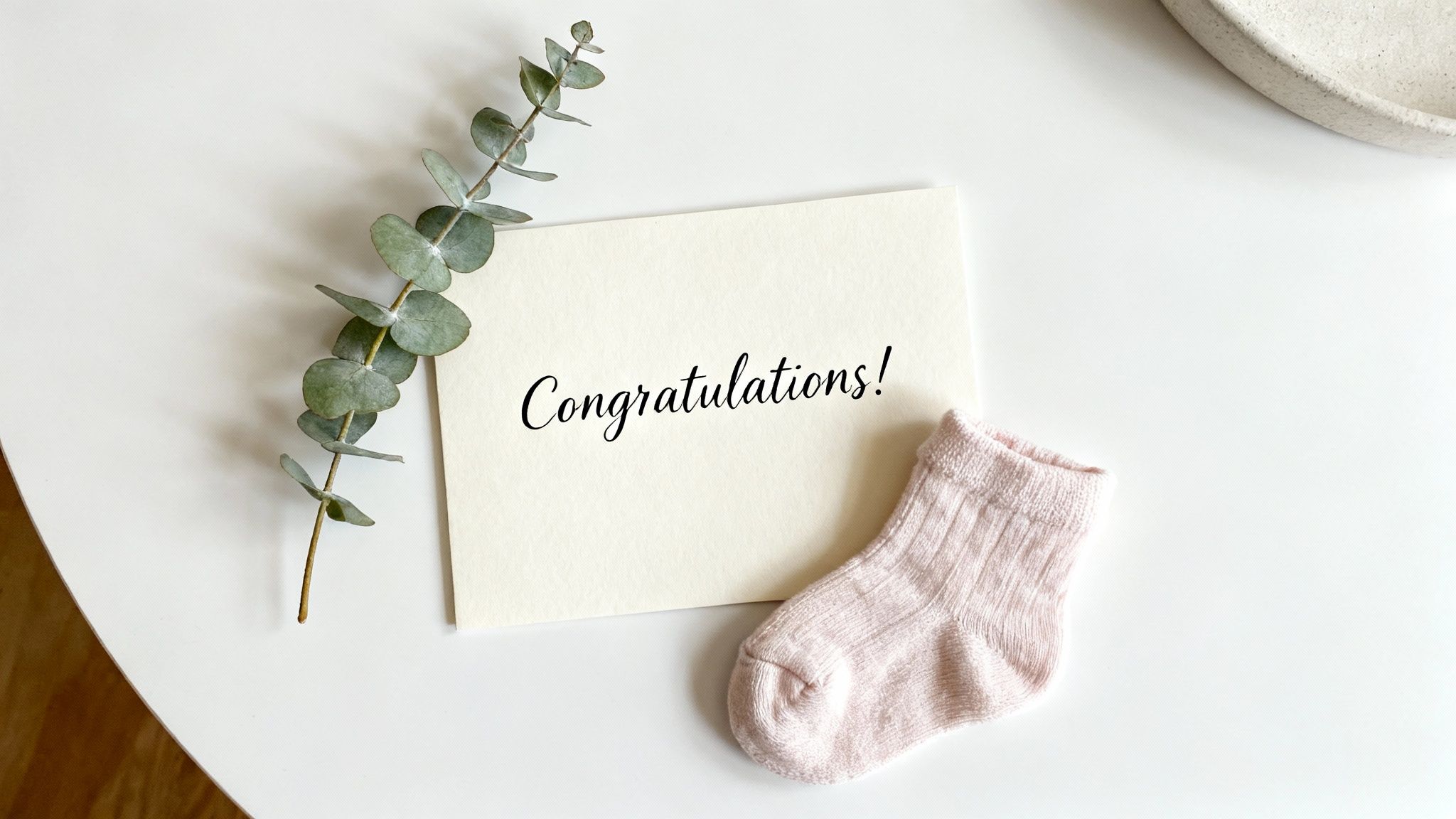 A 'Congratulations!' card with a eucalyptus branch and a pink baby sock on a white surface.
