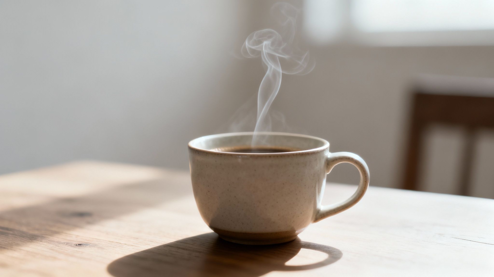 A steaming cup of hot coffee sits on a wooden table, illuminated by sunlight.