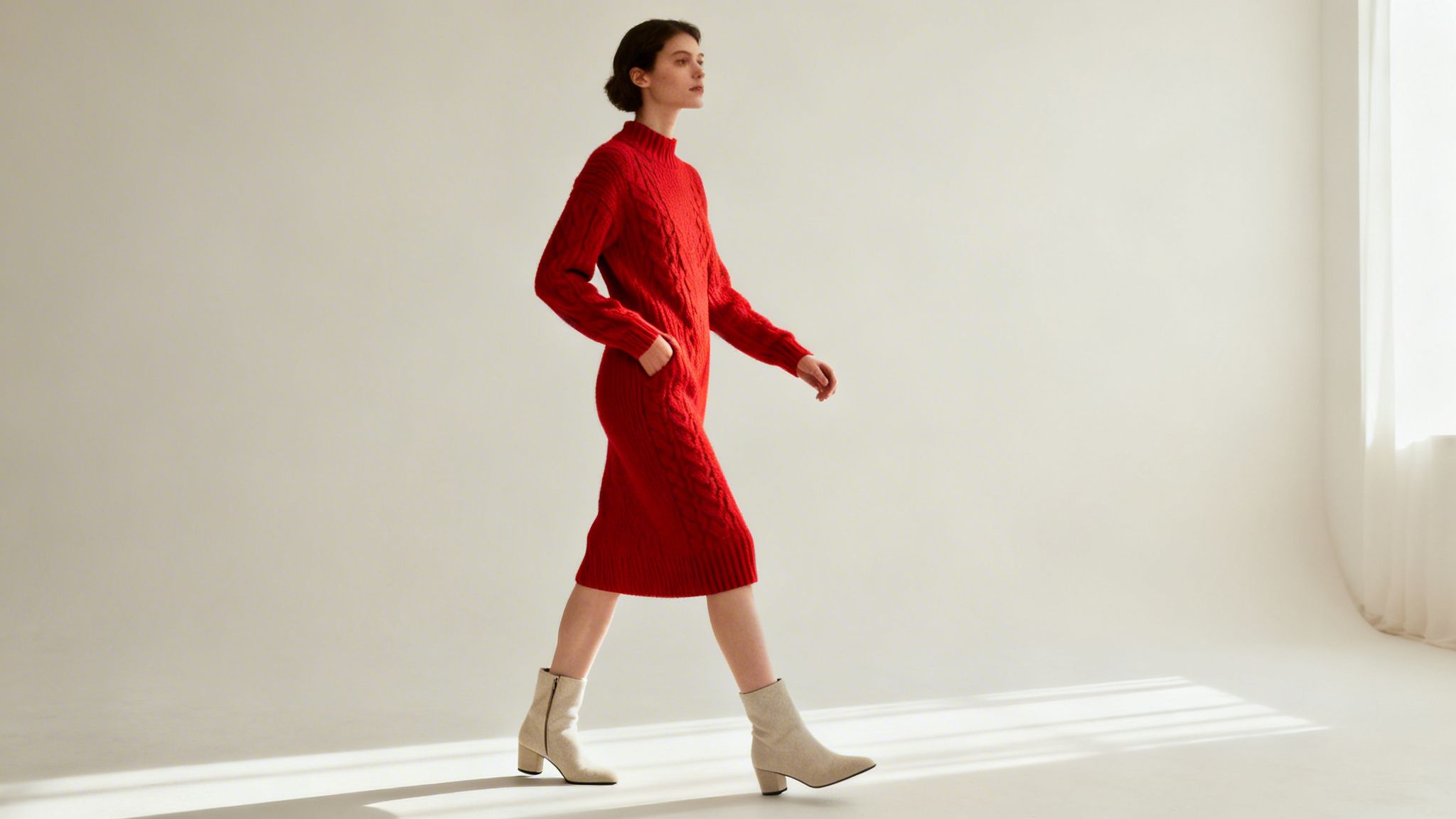 A woman in a red cable-knit dress and beige ankle boots walking in a bright room.
