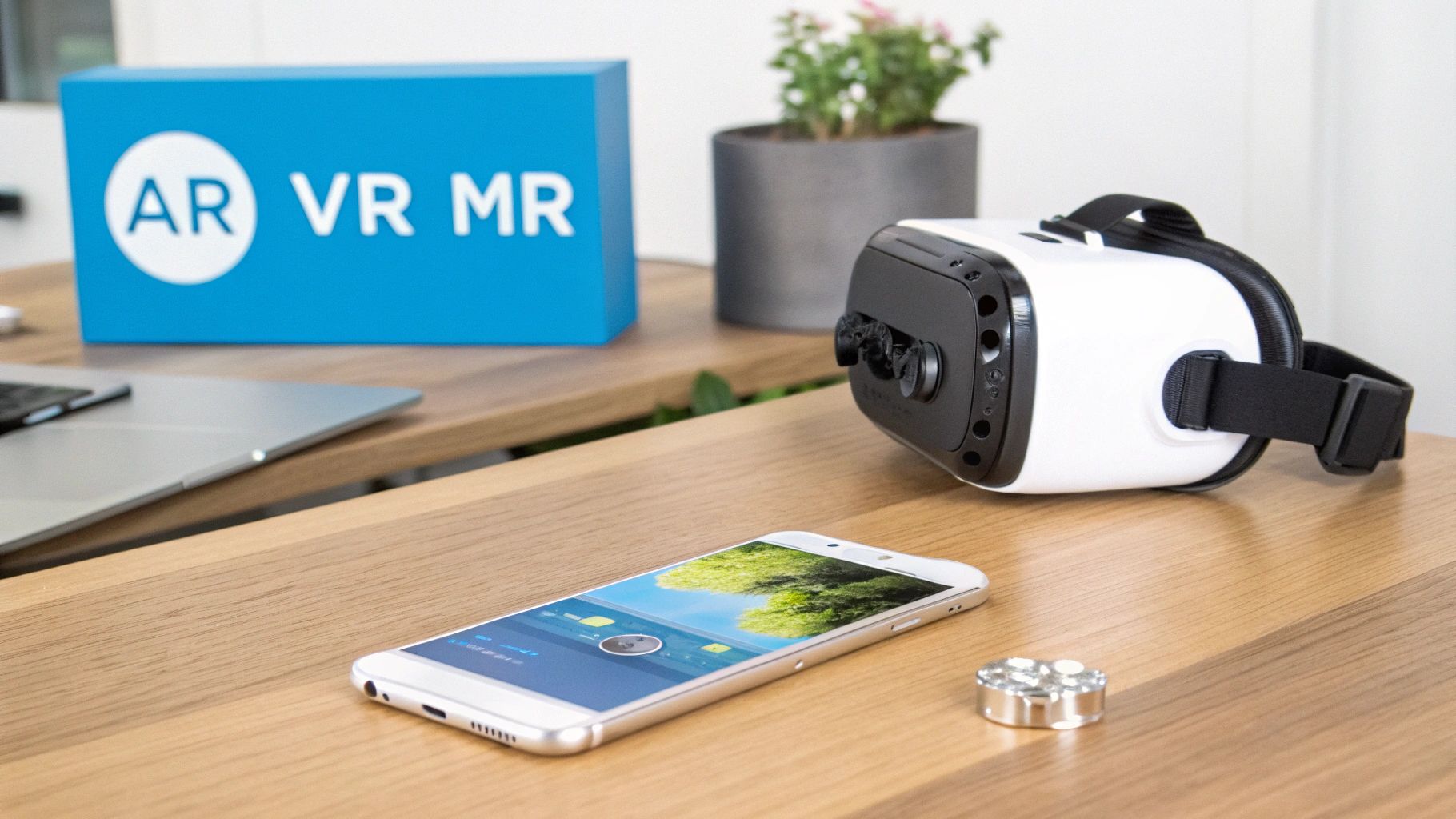 A white VR headset, smartphone, and laptop on a wooden desk with a blue AR VR MR box.