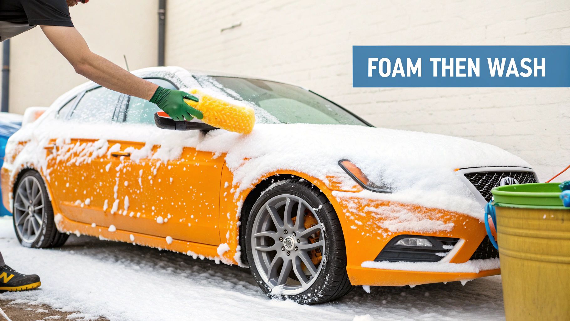 A person uses a yellow mitt to wash an orange car covered in thick white soap foam.