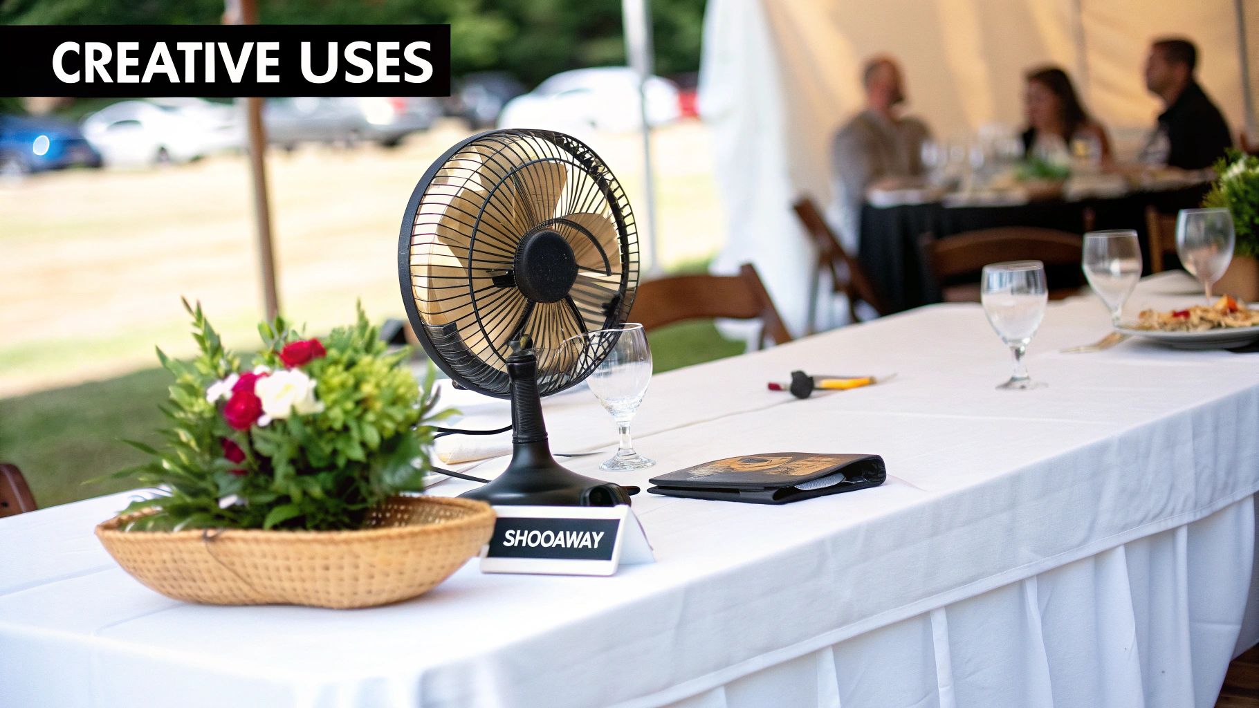 A table at an outdoor event under a tent, featuring a Shooaway fly fan and floral centerpiece.