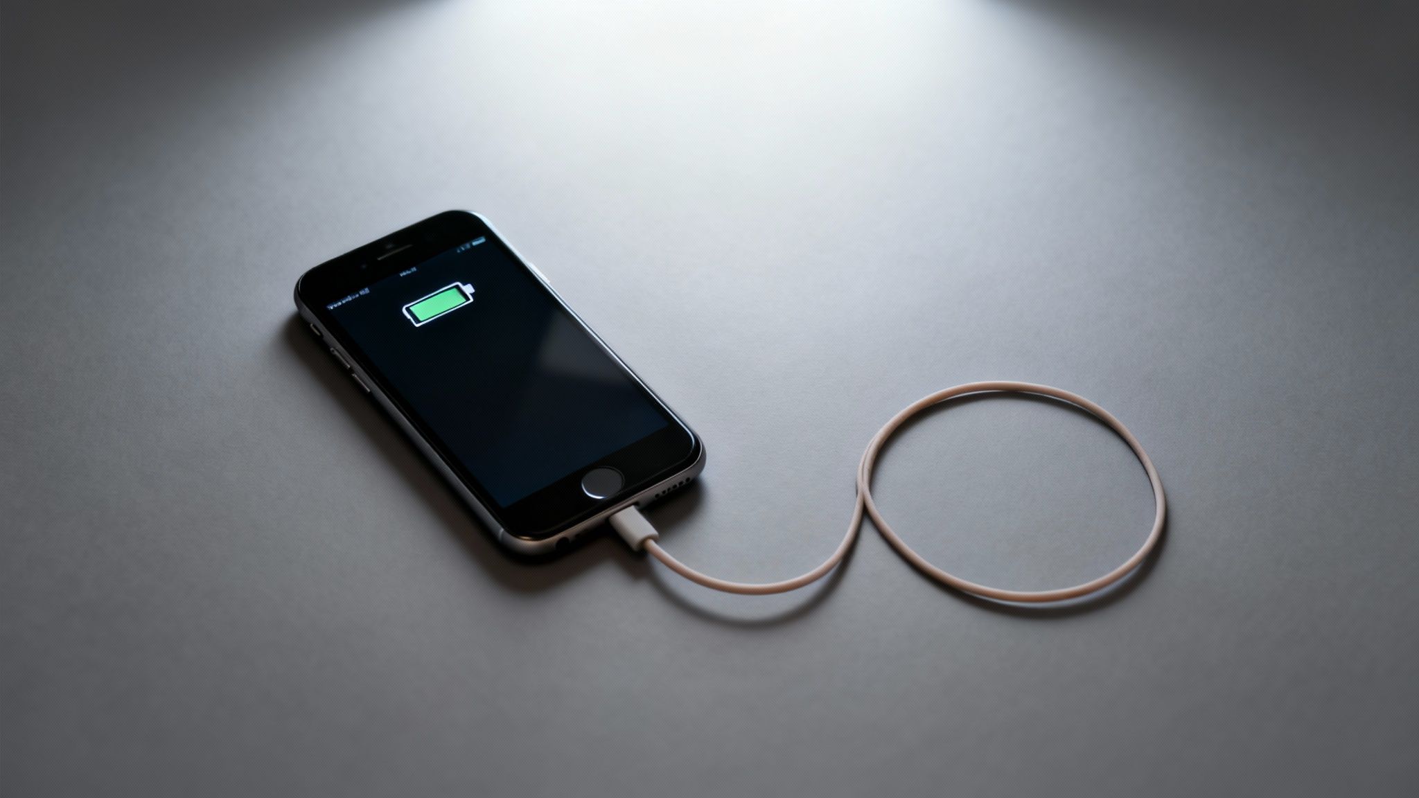 A black iPhone is charging with a white cable, displaying a green battery icon on a dark grey surface.