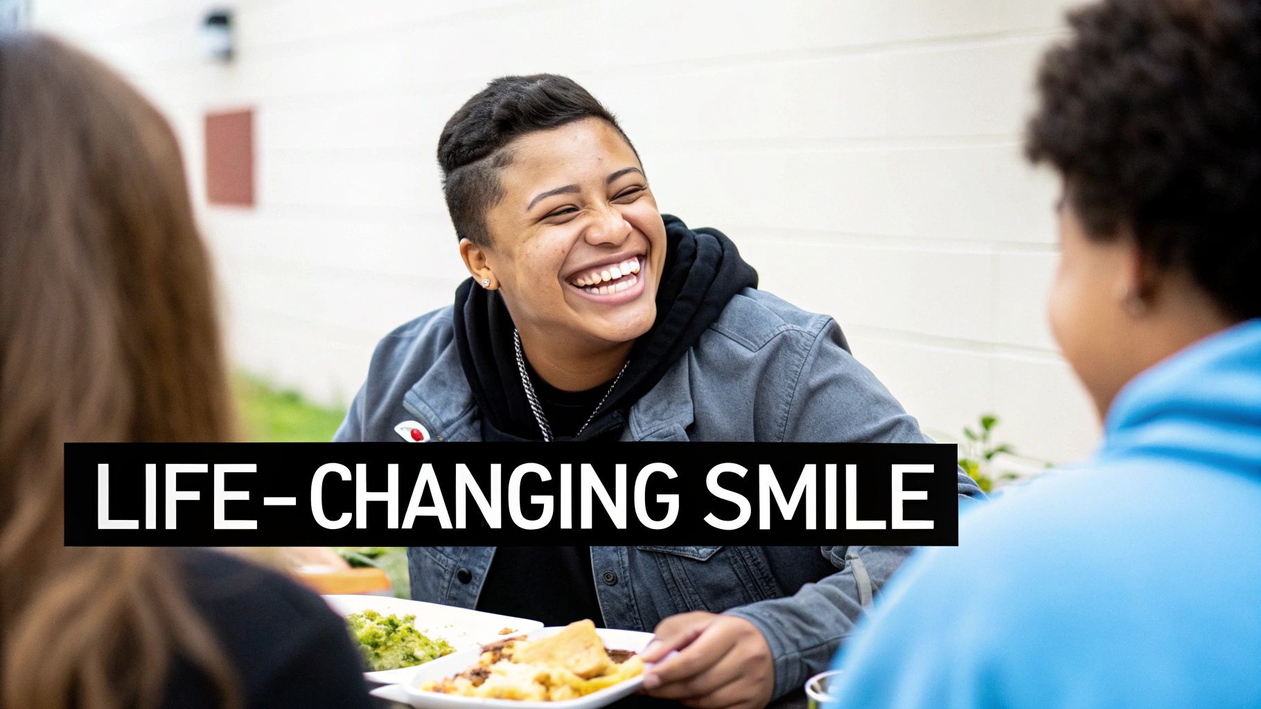 A person with short hair laughs brightly while holding a plate of food, sharing a moment with friends.