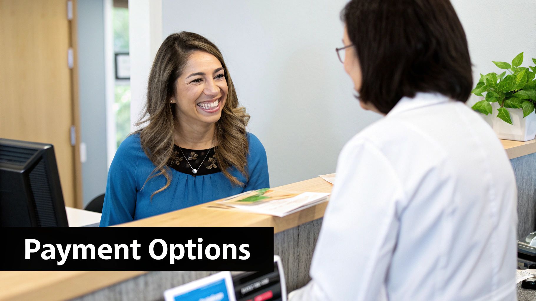 A smiling patient in a blue top discusses payment options with a receptionist at a clinic.