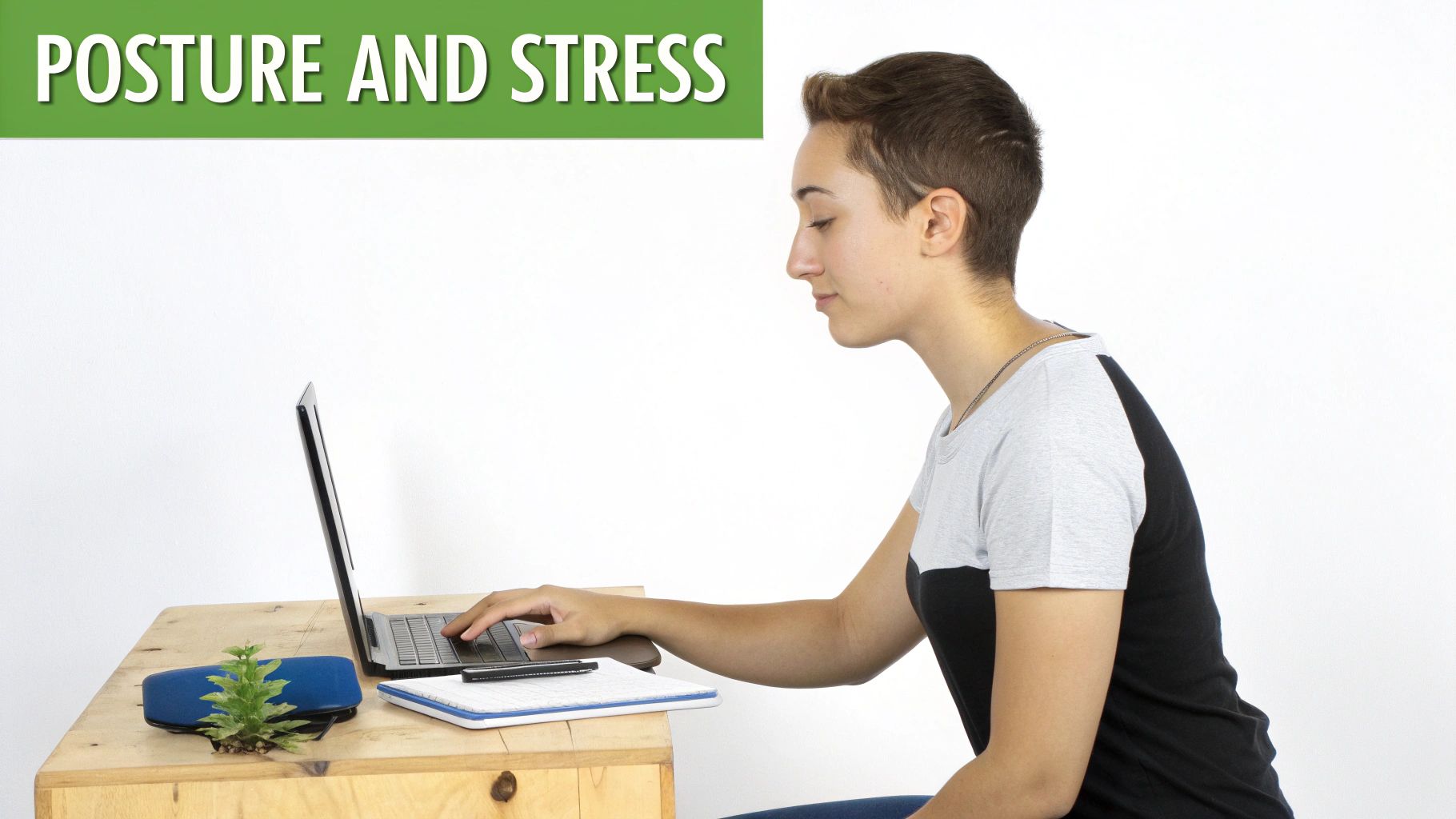 A person with short hair sits at a wooden desk, hunched over a laptop, typing.
