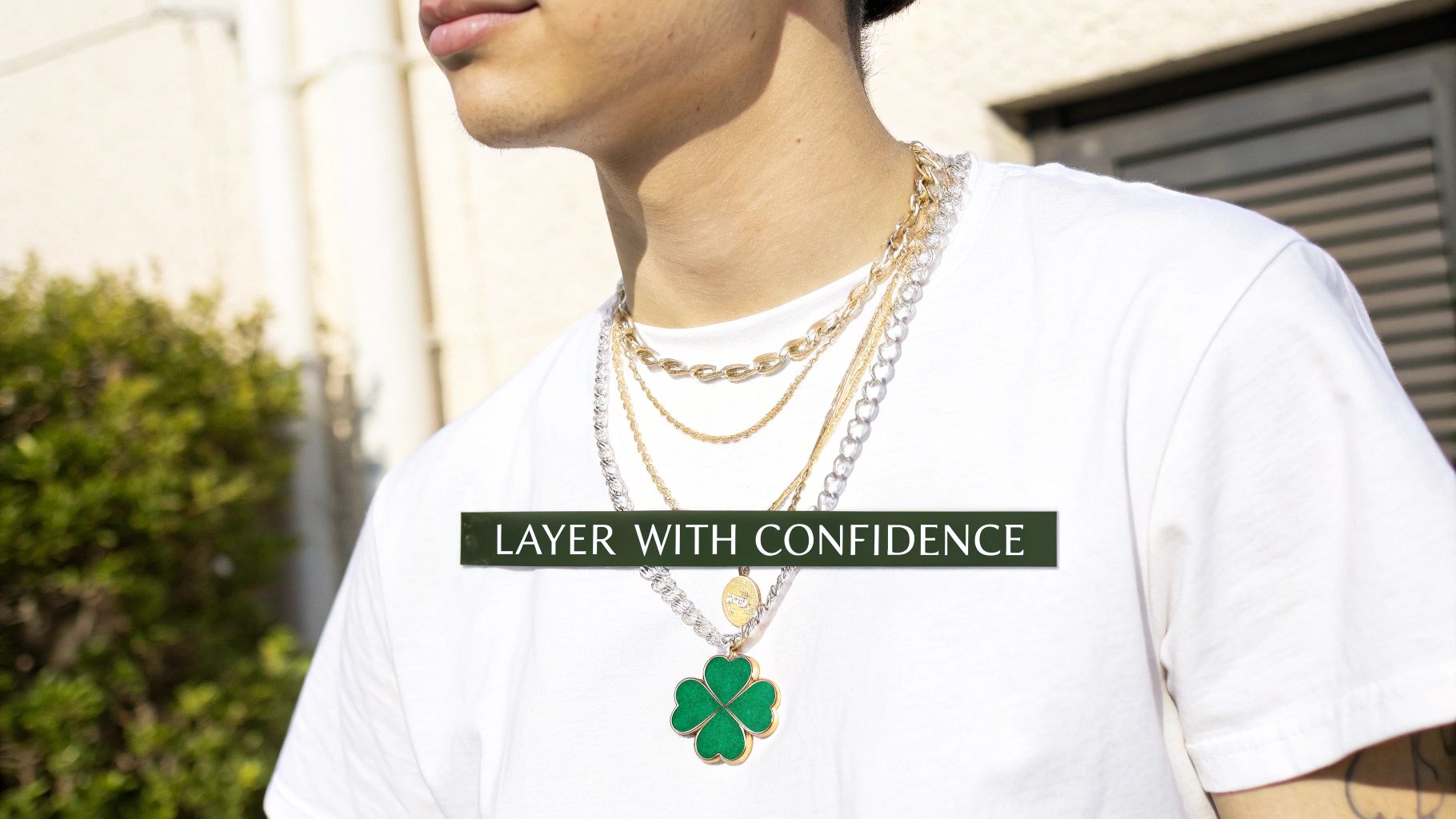 A person wearing a white t-shirt, layered gold and silver chains, and a green four-leaf clover necklace.
