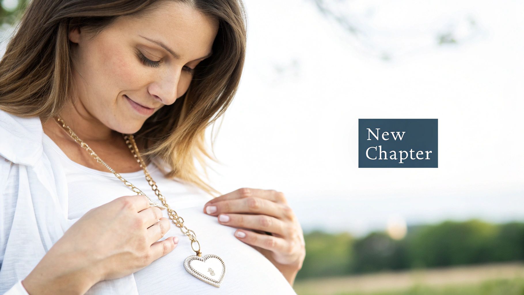 A smiling pregnant woman gently holds her belly, wearing a prominent gold heart pendant necklace.