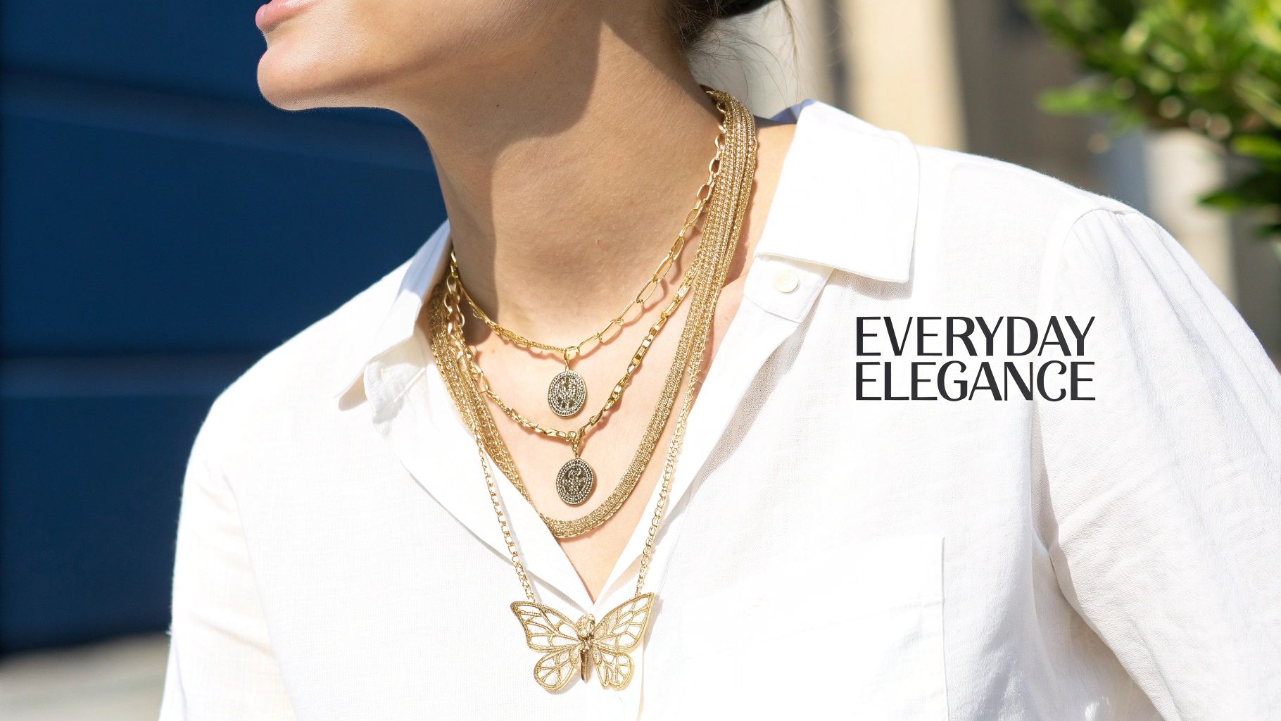 Stylish woman wearing layered gold necklaces, featuring a large butterfly pendant, on a white shirt.
