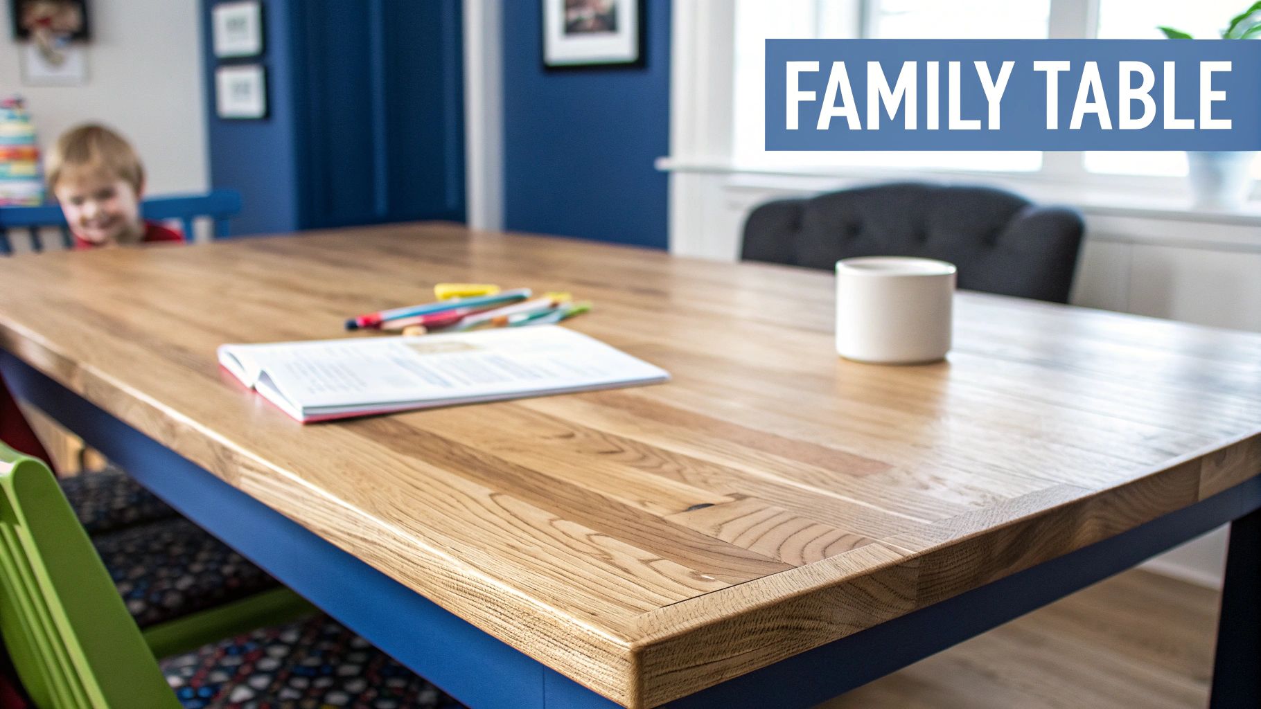 A light wooden family table with a blue base, a book, pencils, and a smiling child.