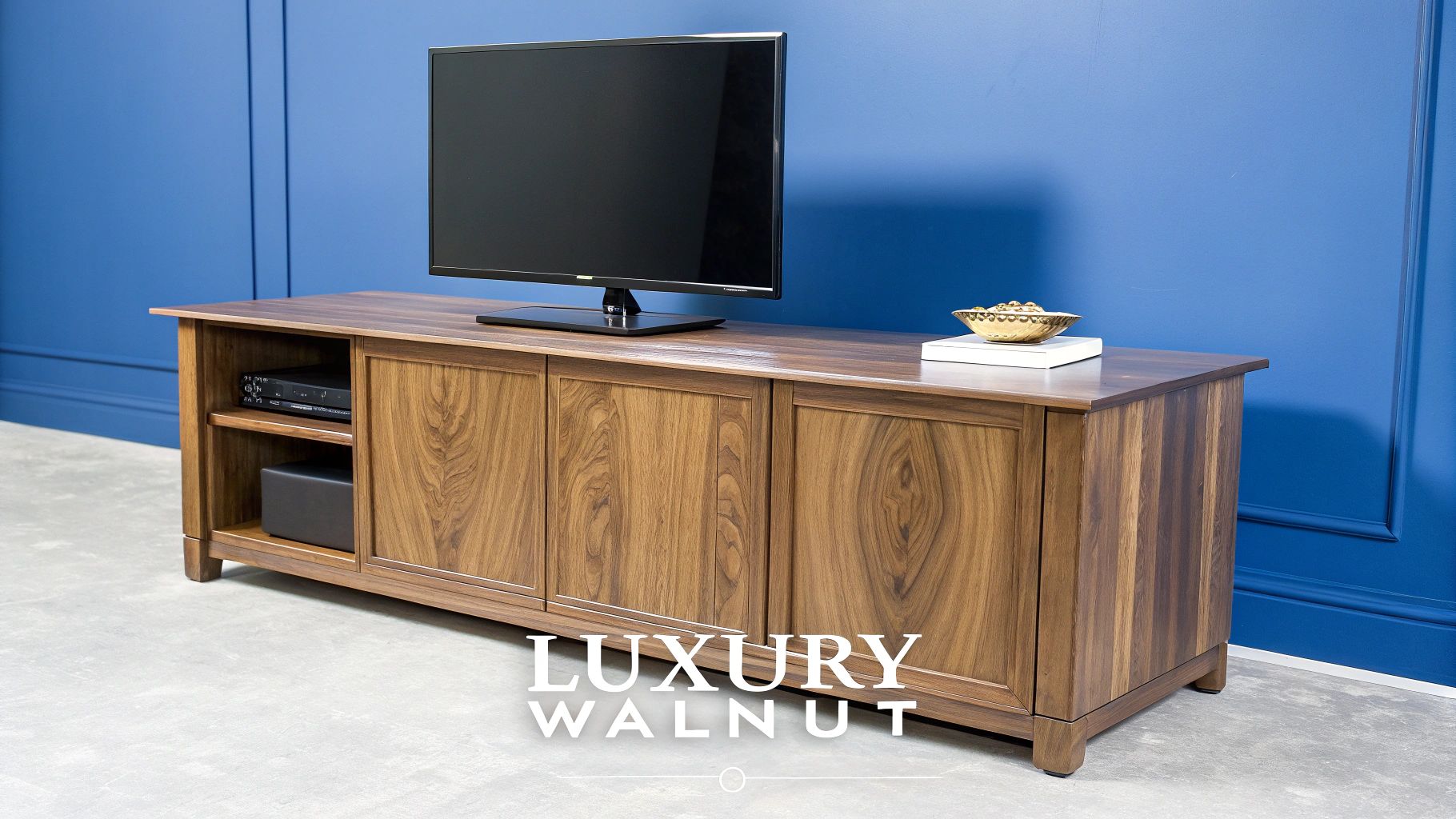A luxurious walnut wood TV stand with a television, media devices, and decorative bowl against a blue wall.