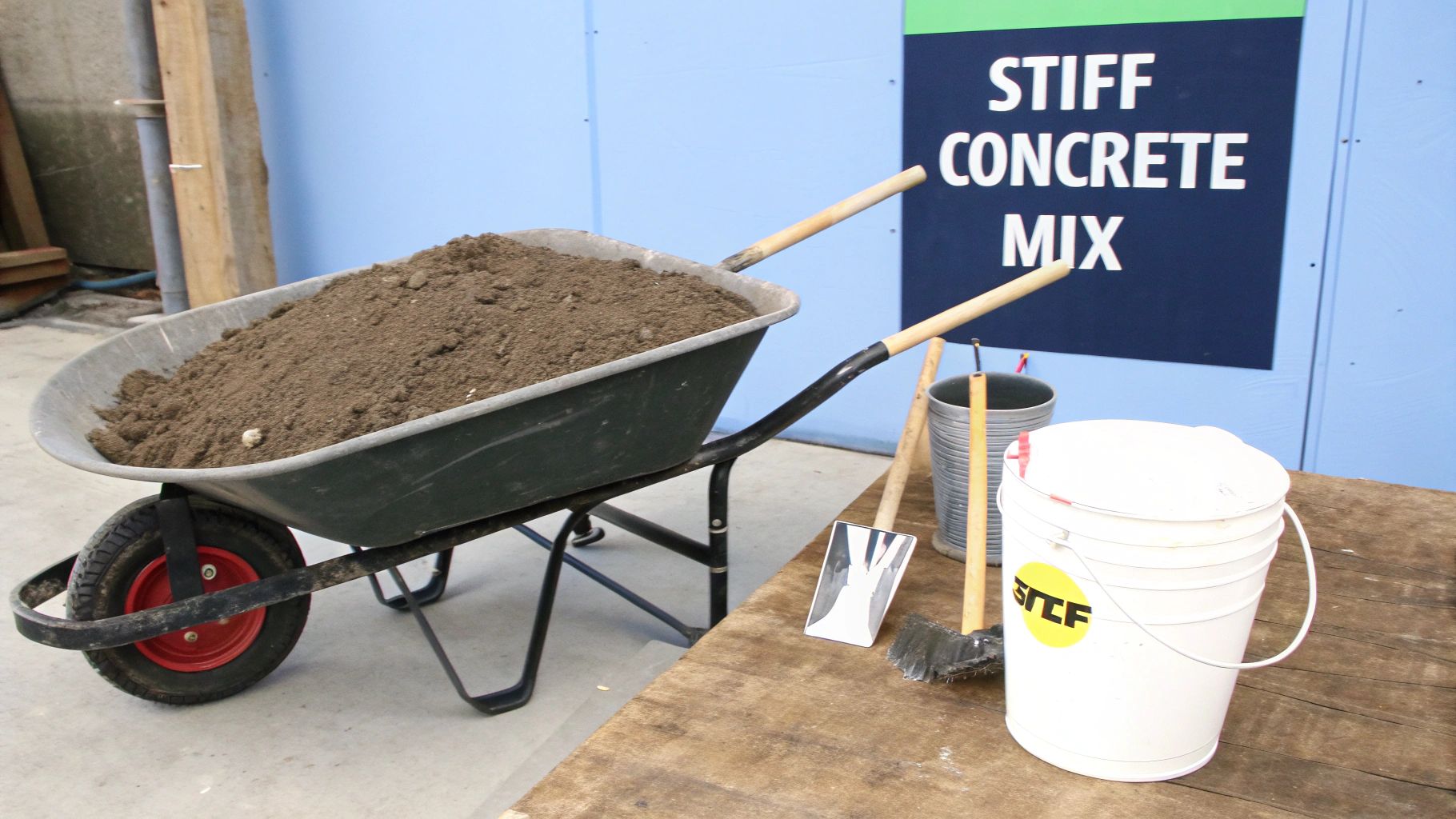 A wheelbarrow filled with soil, construction tools, buckets, and a "STIFF CONCRETE MIX" sign.