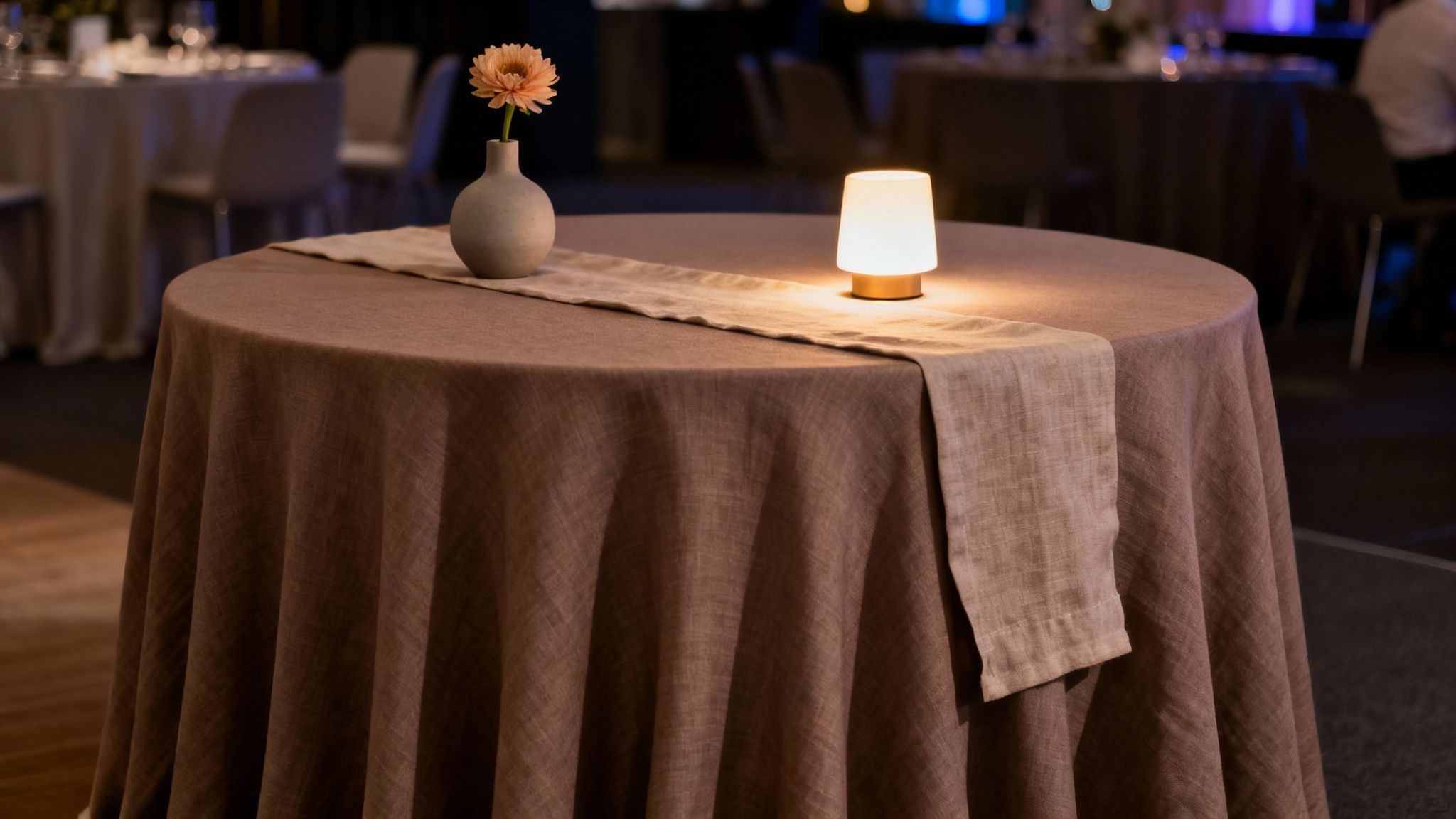 Een ronde tafel met bruin tafelkleed, beige tafelloper, vaas met oranje bloem en sfeervolle tafellamp in een evenementenruimte.