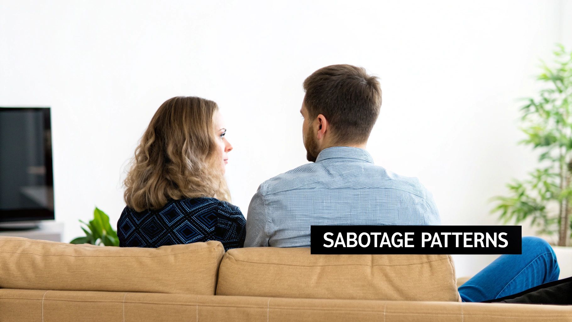 A couple sits on a couch, the woman looking at the man, discussing sabotage patterns.