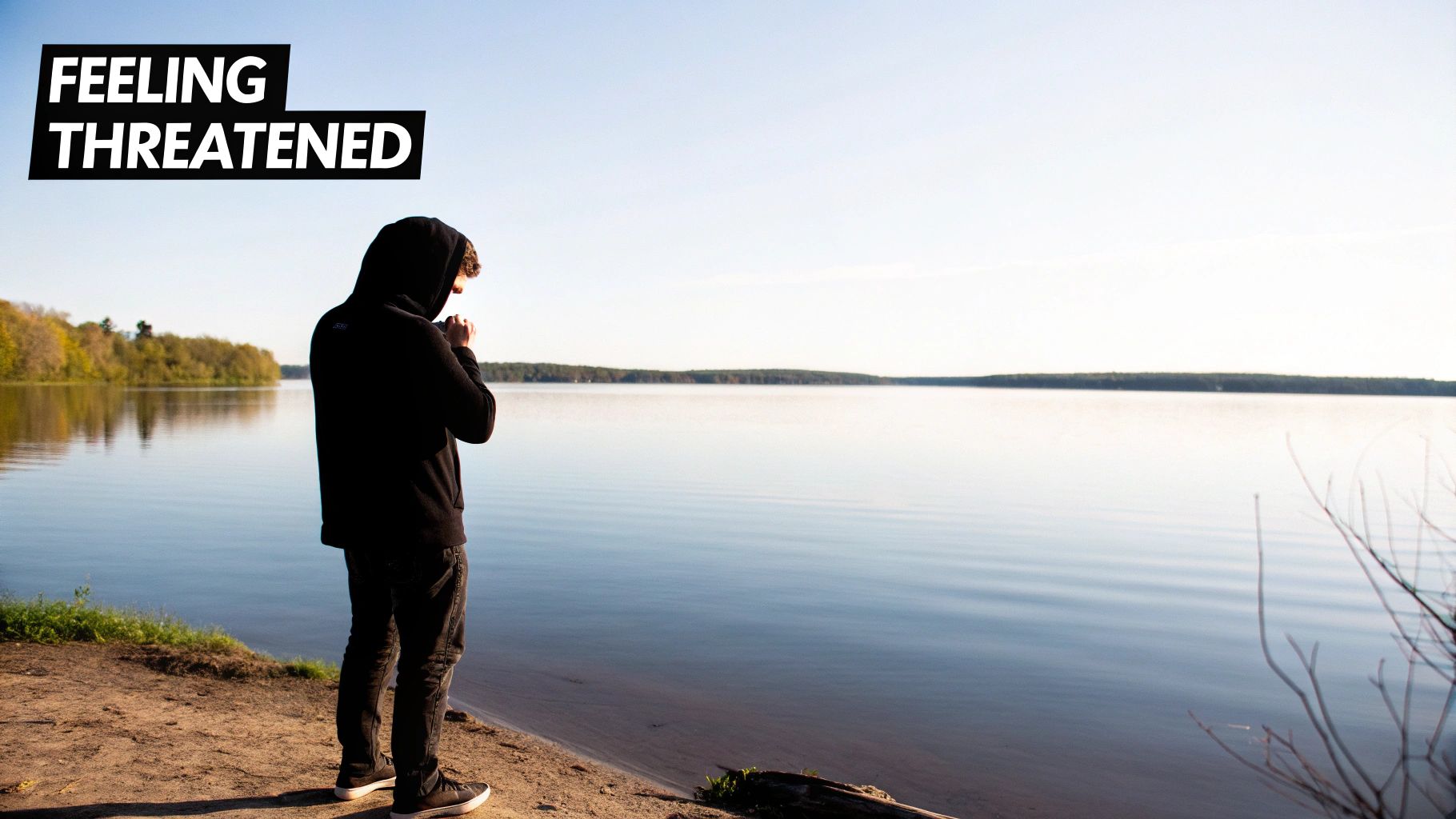A solitary person in a black hoodie stands by a calm lake, feeling threatened.