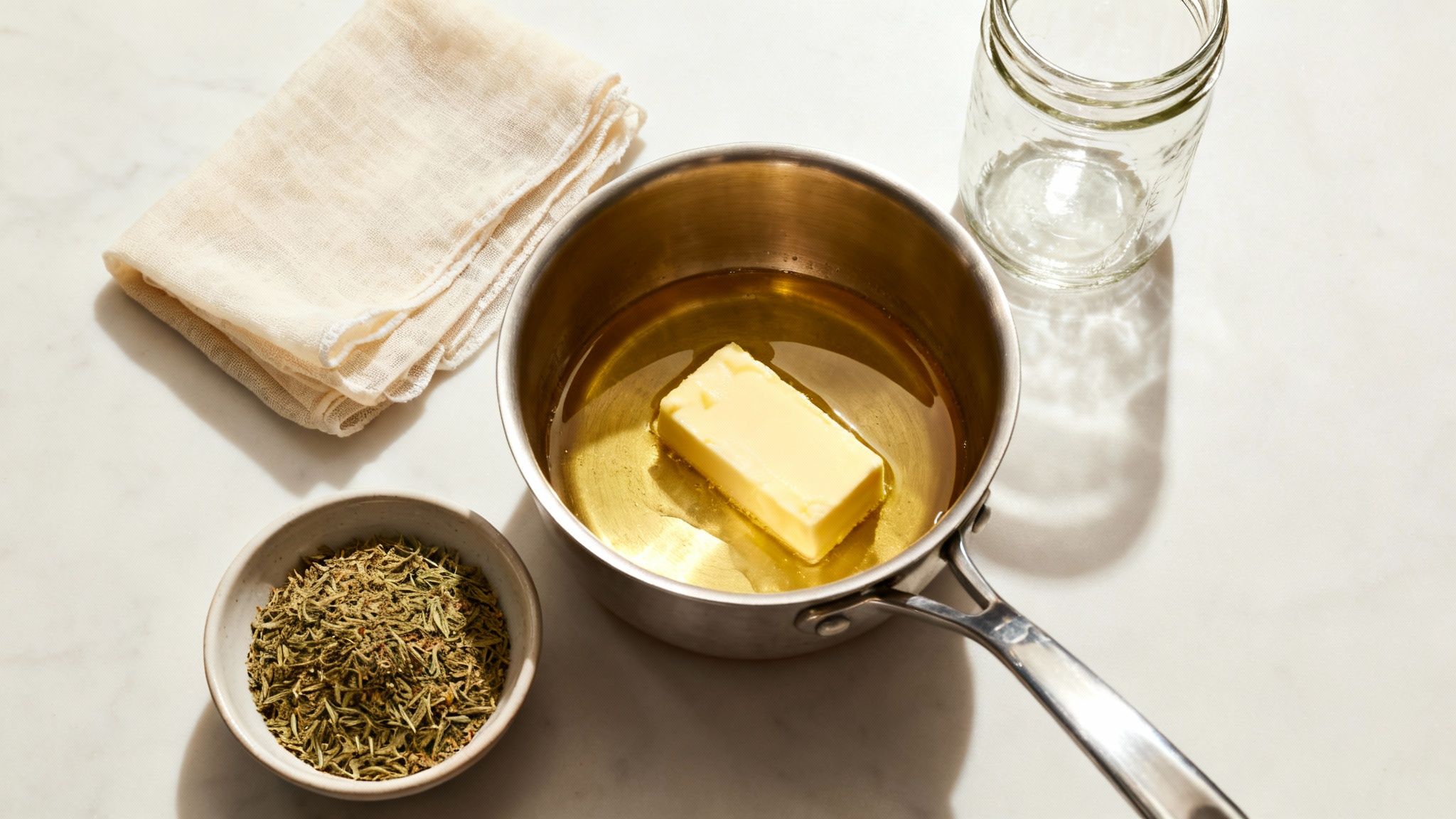 Ein Topf mit Butter in Öl, getrocknete Kräuter, Käsetuch und Glas auf einer Marmoroberfläche.