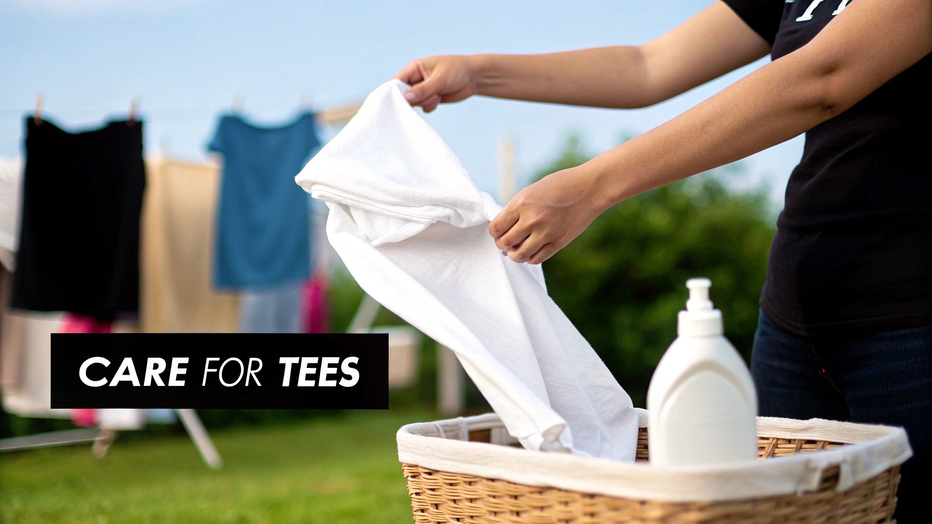 A person's hands are placing a white t-shirt into a laundry basket outdoors, with clothes drying.
