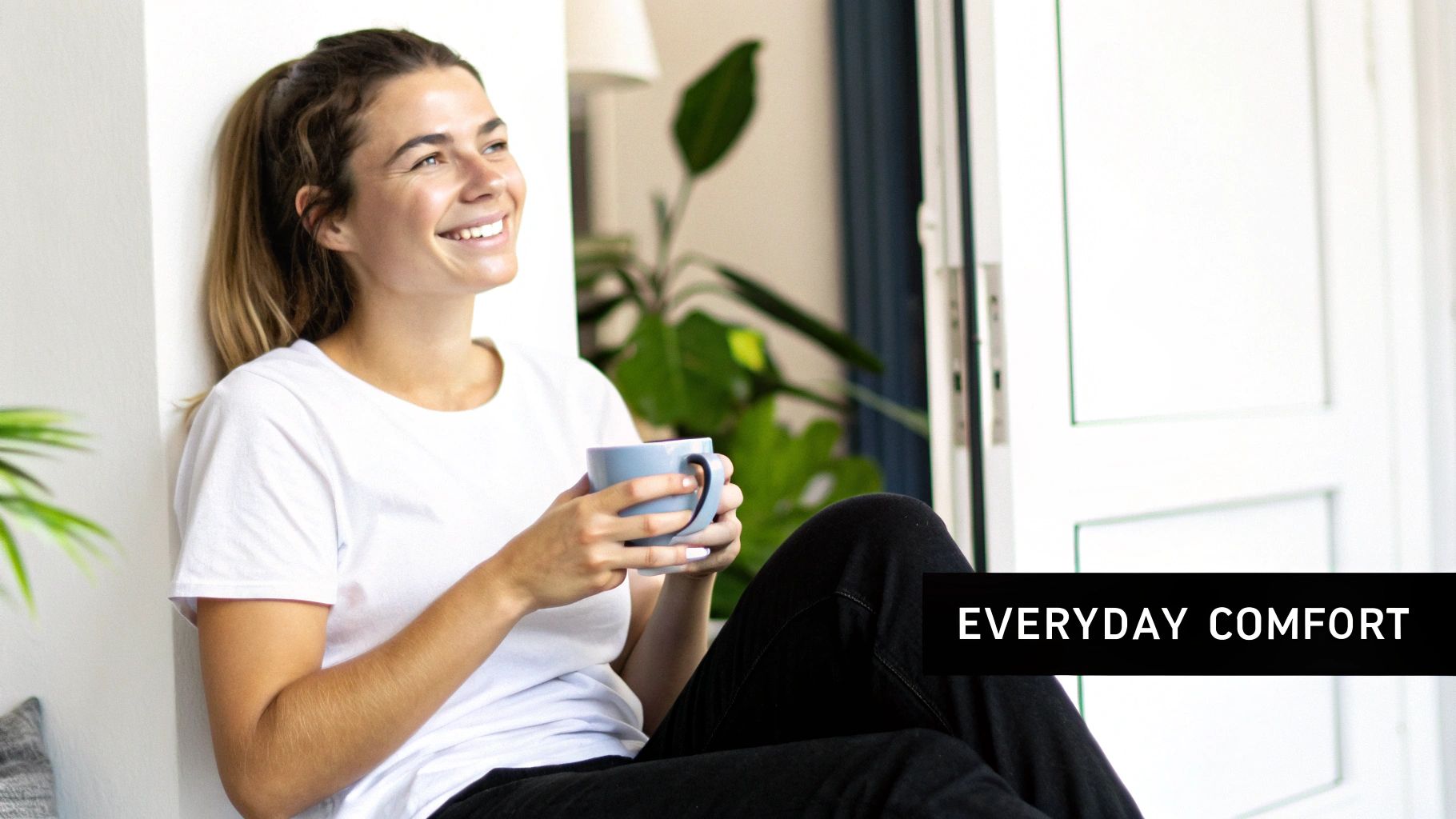 Smiling woman in a white t-shirt enjoying a warm drink in a comfortable home setting.