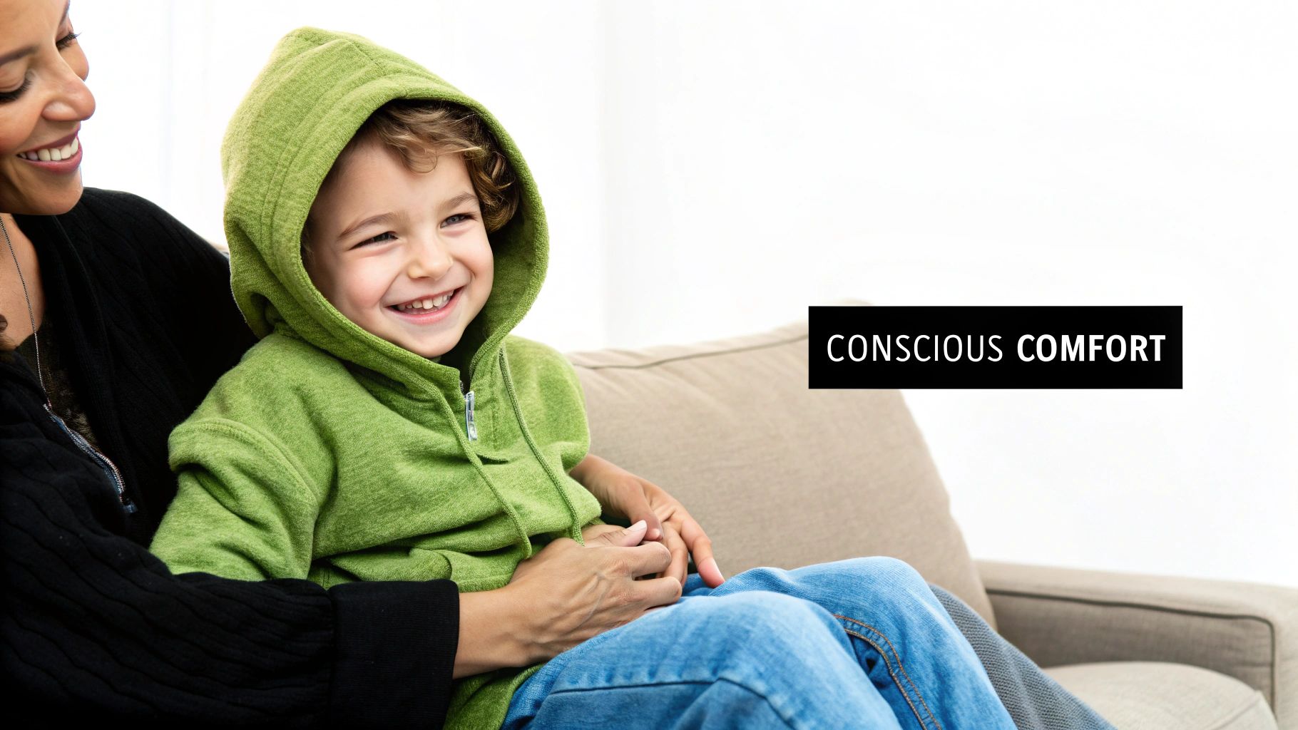 A smiling woman holding a happy child in a green hoodie on a beige couch.