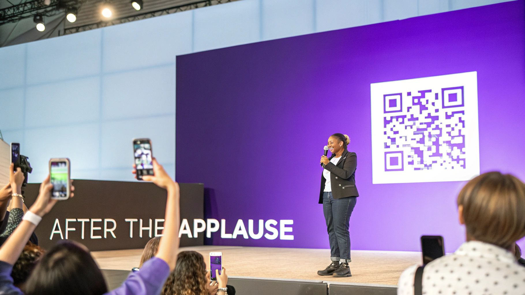 A smiling speaker presents on stage next to a large QR code, with audience members recording.