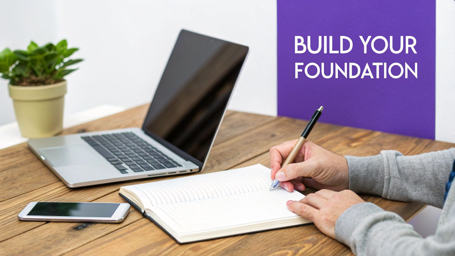Person writing in a notebook on a wooden desk with a laptop and smartphone, text says 'BUILD YOUR FOUNDATION'.