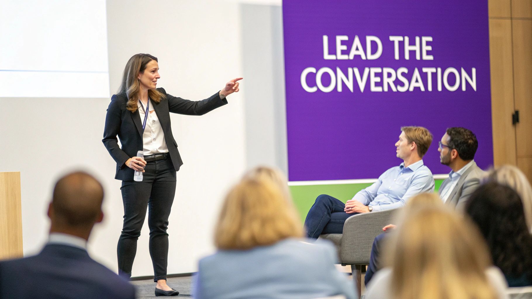 A moderator leading a panel discussion with engaged panelists and an audience in the background.