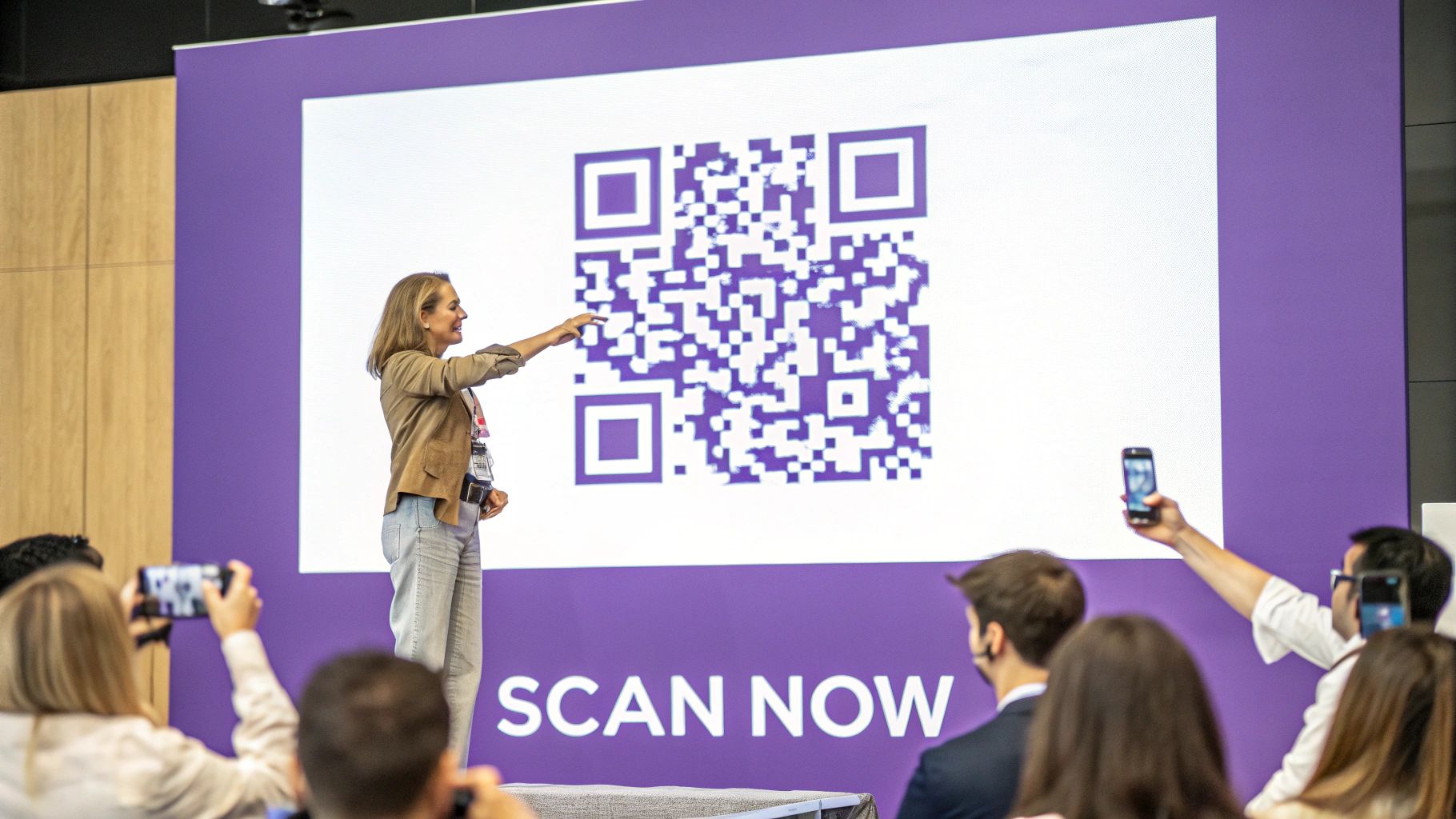 A woman points at a large purple QR code on a screen during a presentation, with audience members scanning it.